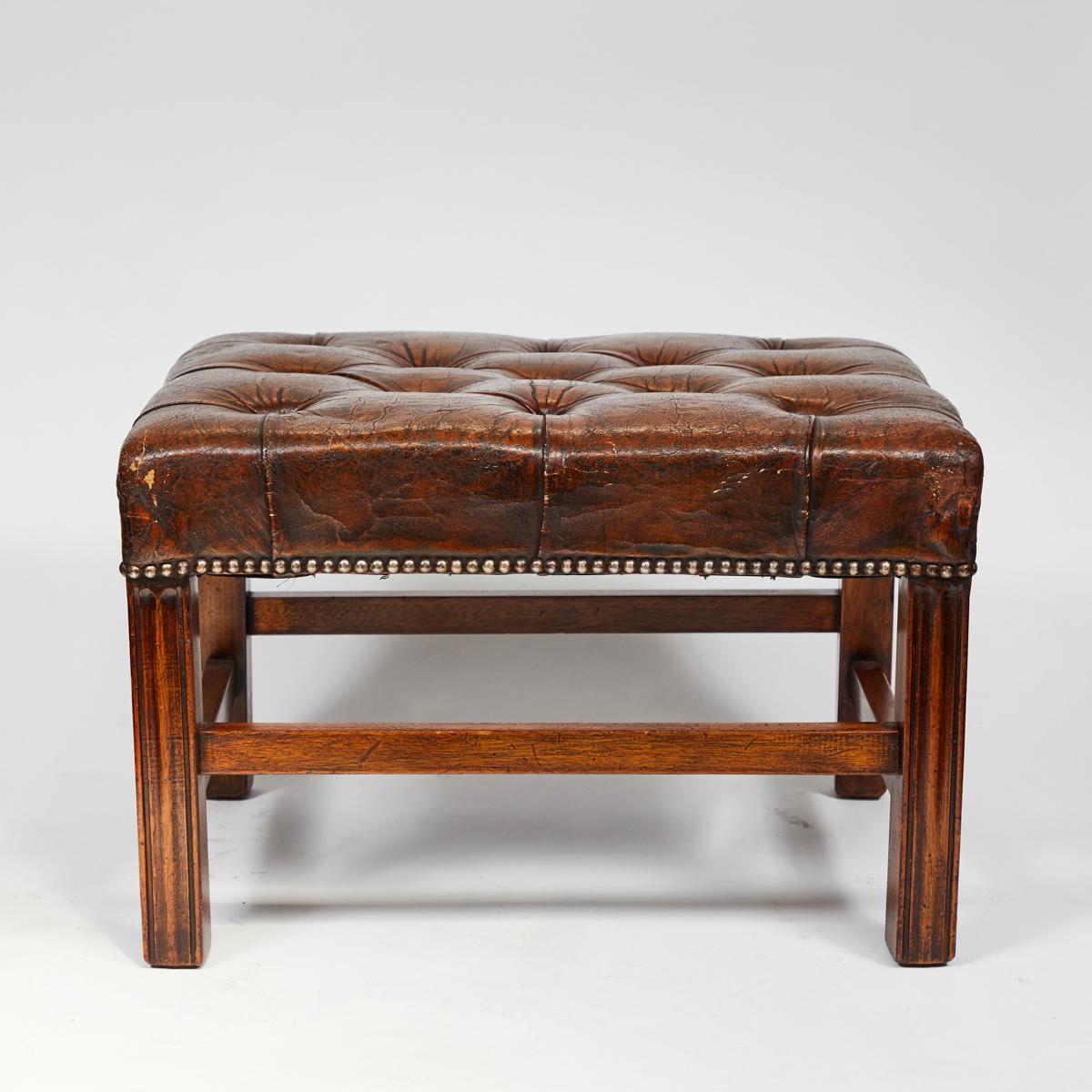 Early 20th-century English tufted upholstered leather footstool or bench. This stool has acquired an attractive patina over the course of its life. The block-style legs feature carved vertical grooves, and the gently-worn upholstery is affixed at