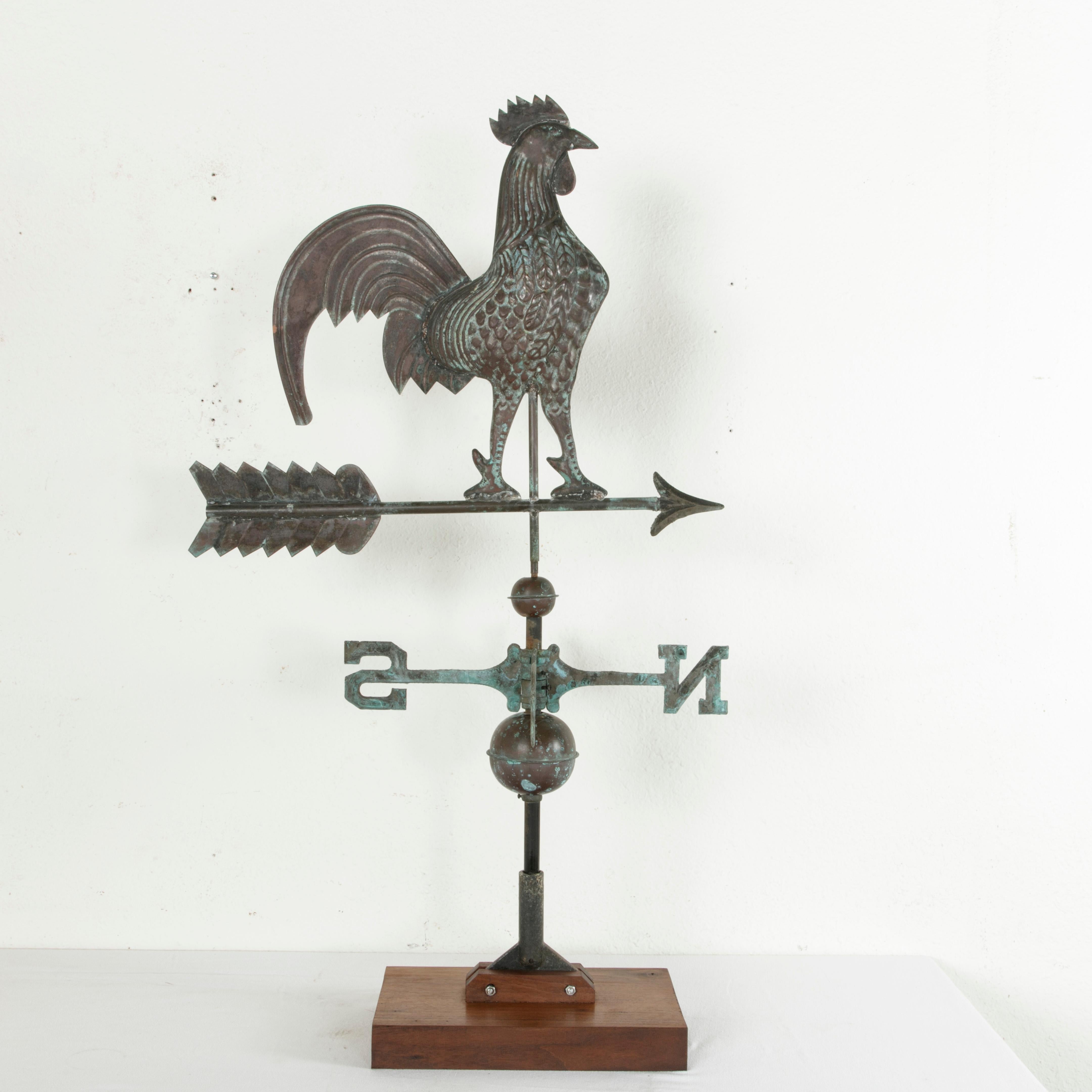 Found in France, this early 20th century patinated copper weather vane features a rooster perched on top of an arrow. Below are arms with the letters NESW indicating the cardinal directions in English. A spinning ball is above and below the arms.