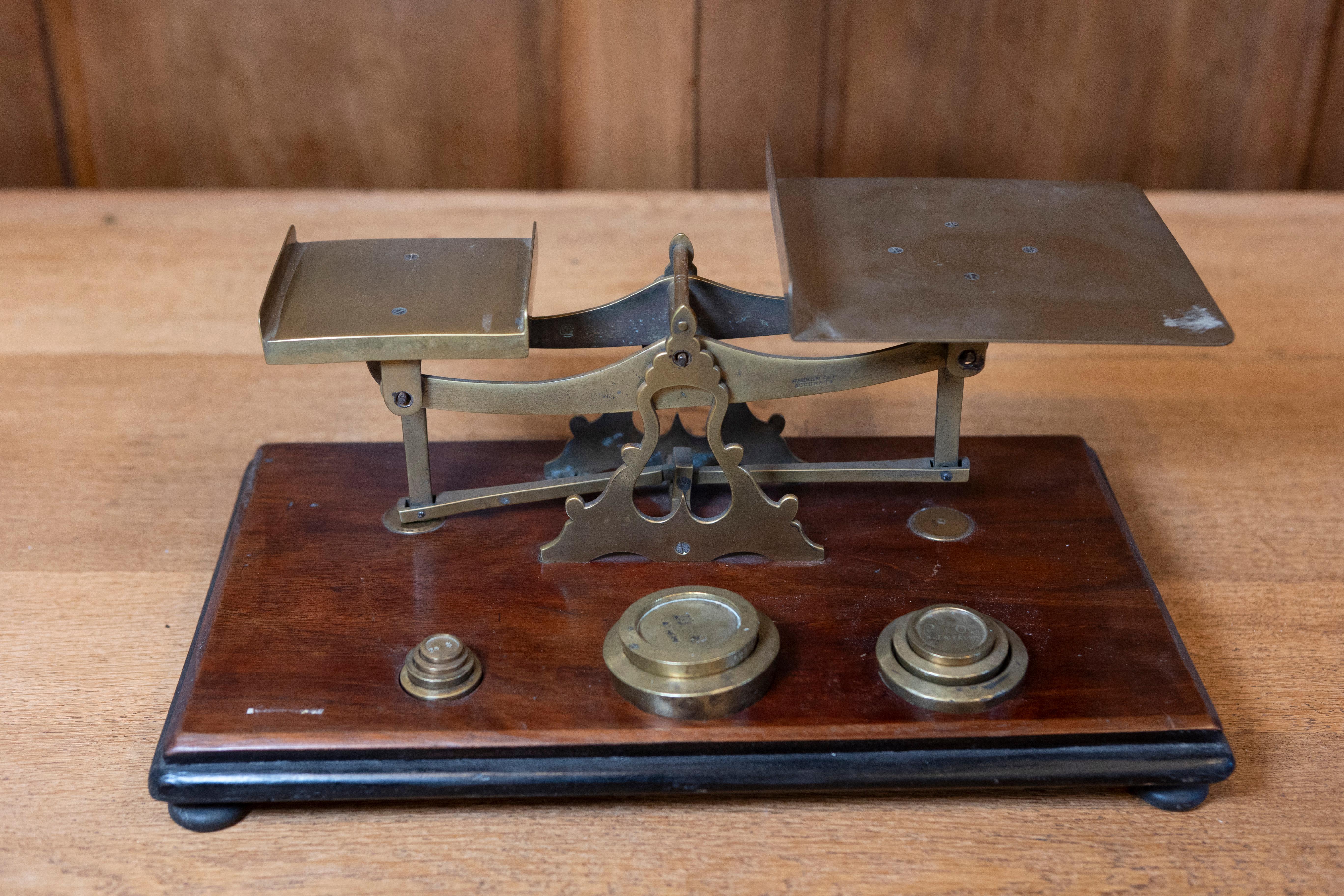 This finely crafted Early 20th Century English postal scale is a striking example of the functional yet elegant design typical of the period. The scale is mounted on a polished wooden base, likely made from mahogany or walnut, known for its rich