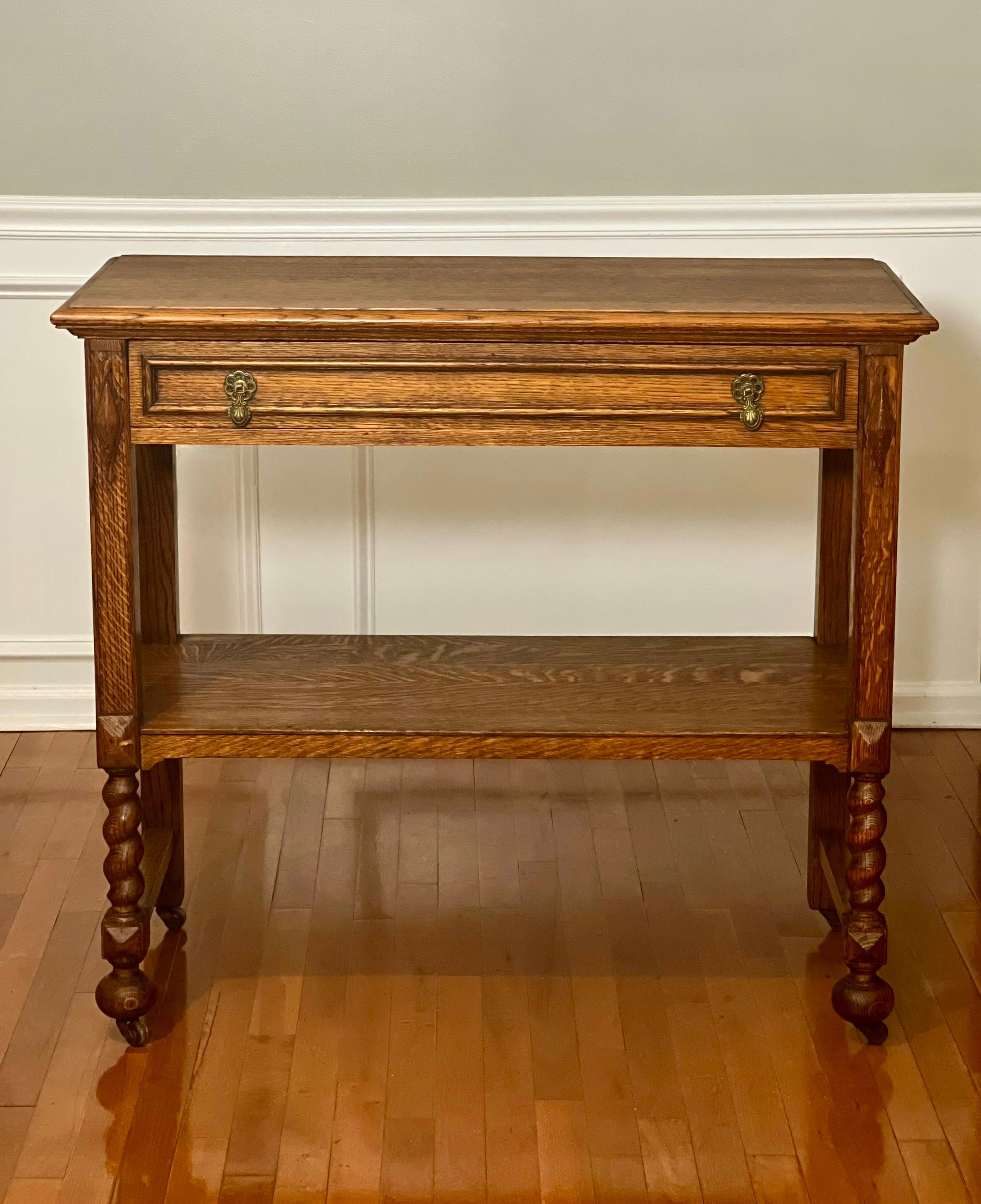 English quarter sawn oak two-tier server or console on casters, c. 1910.

Handsome server featuring a single dovetailed drawer with teardrop brass pulls and barley twist front supports. Carved, high relief diamond motifs flank the raised panel