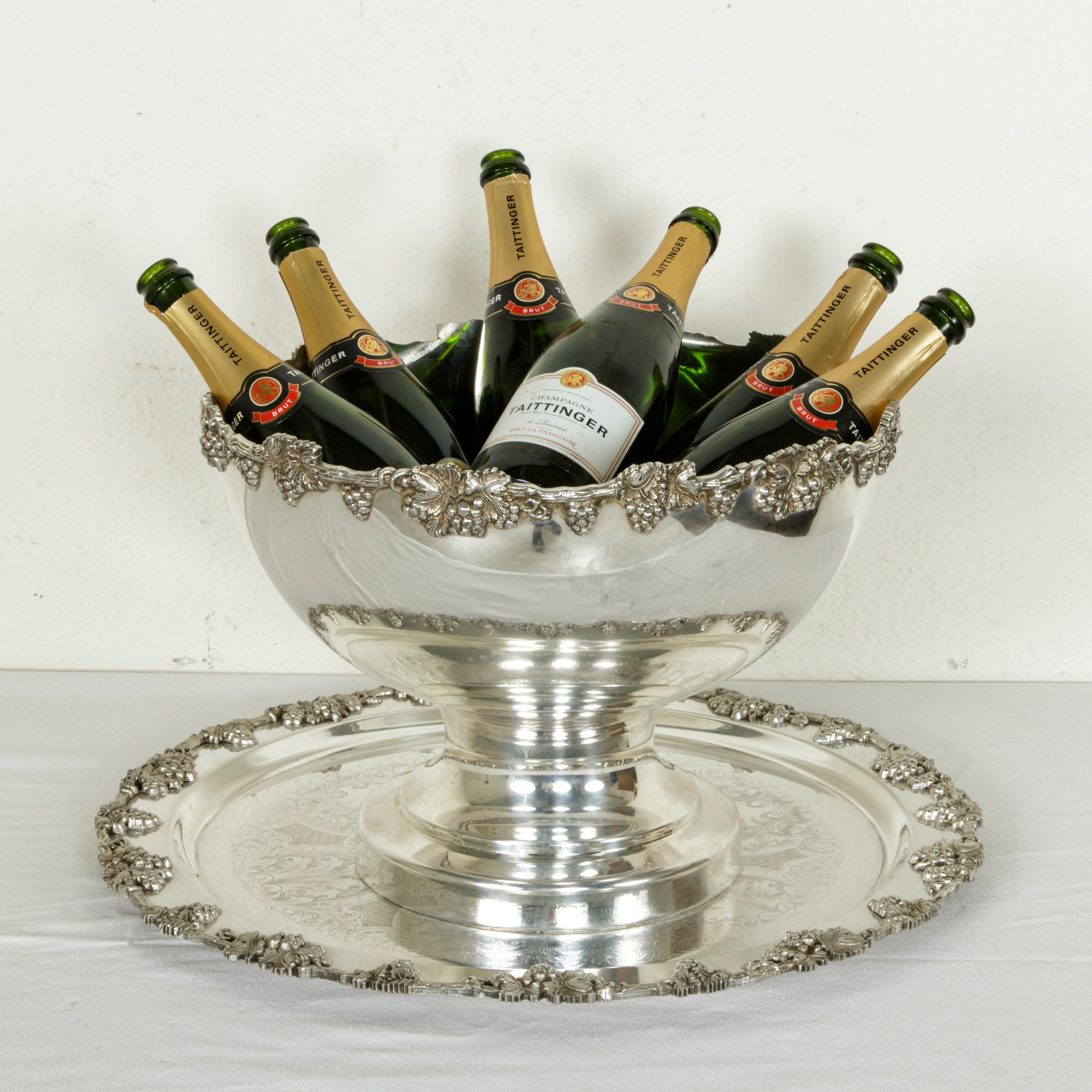 This large early 20th century English silver plate footed hotel champagne bucket holds up to six bottles and is accompanied by a matching tray. The bucket and tray feature borders of grapes and grape vines. The tray measures 21 inches in diameter