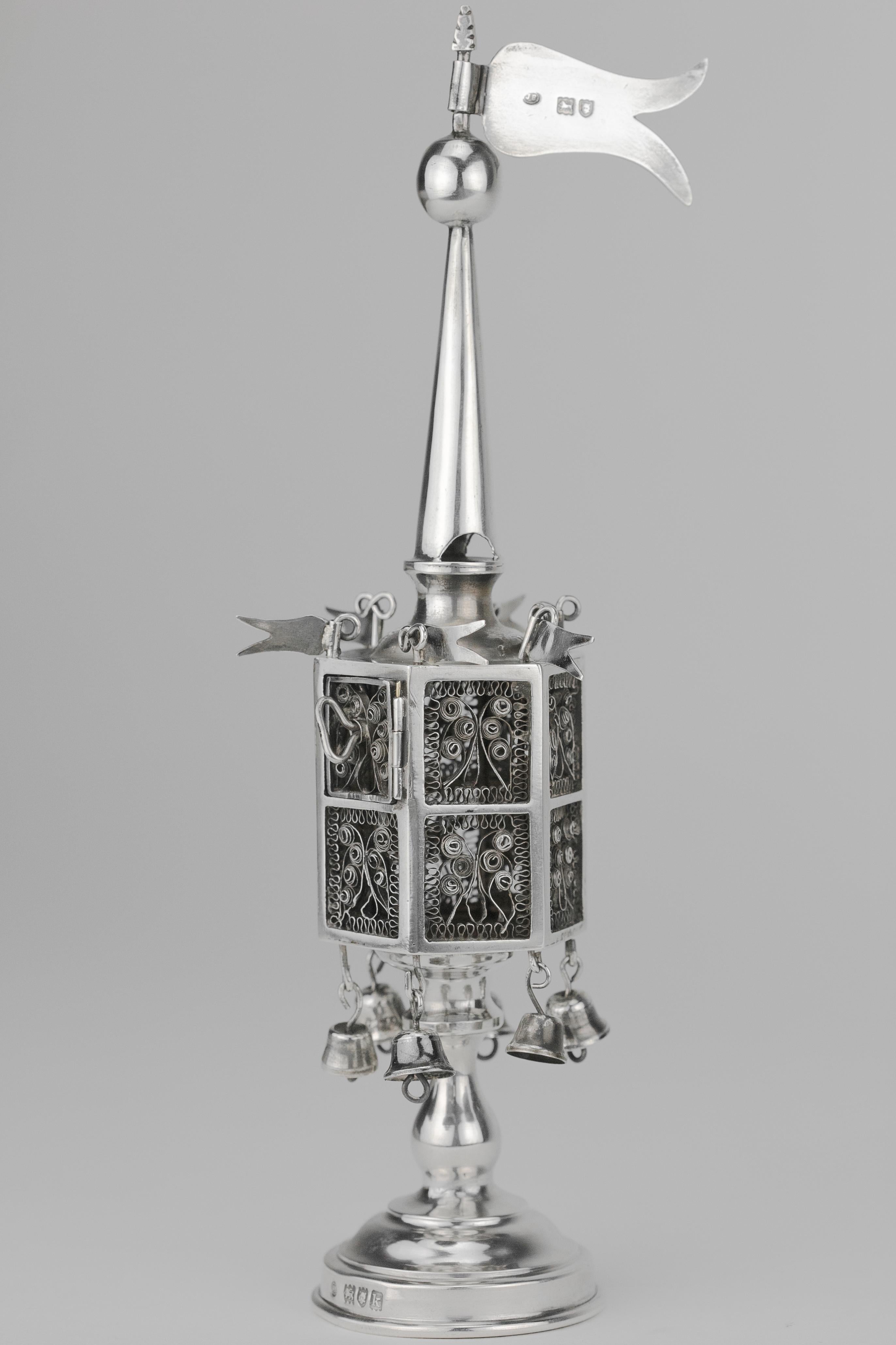 Sterling silver spice tower, by Jacob Fenigstein, Birmingham, England, 1909.
The hexagonal, silver filigree openwork body is fitted with hinged door, six hanging bells and six pennants, topped by a spire, knop and pennant and standing on round base.