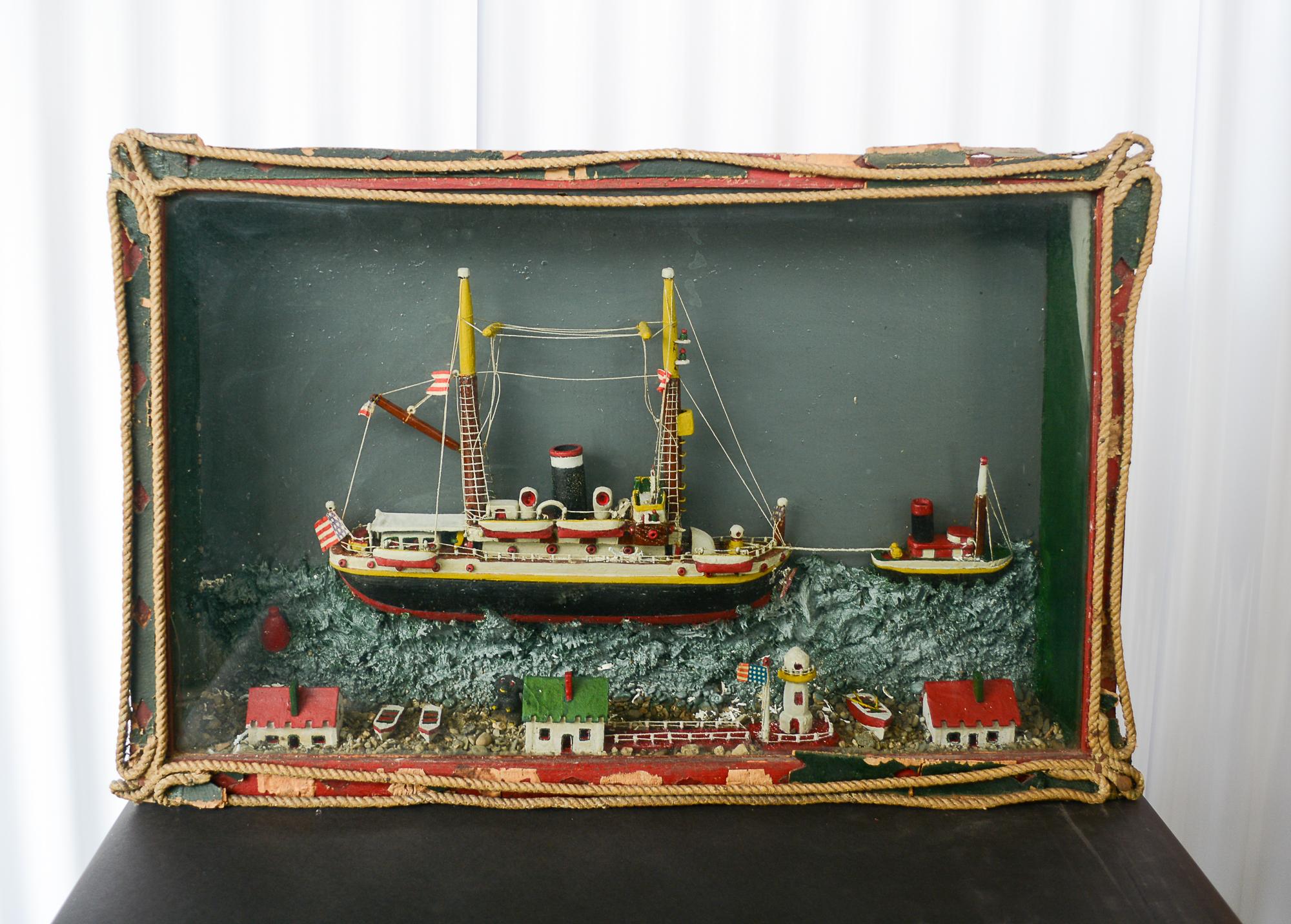 Folk art diorama of a ship being towed by a tug boat. At the bottom of the diorama is a representation of the shore with buildings and a lighthouse. The glass is earlier wavy glass and the back appears to be one solid wide board. The outside below