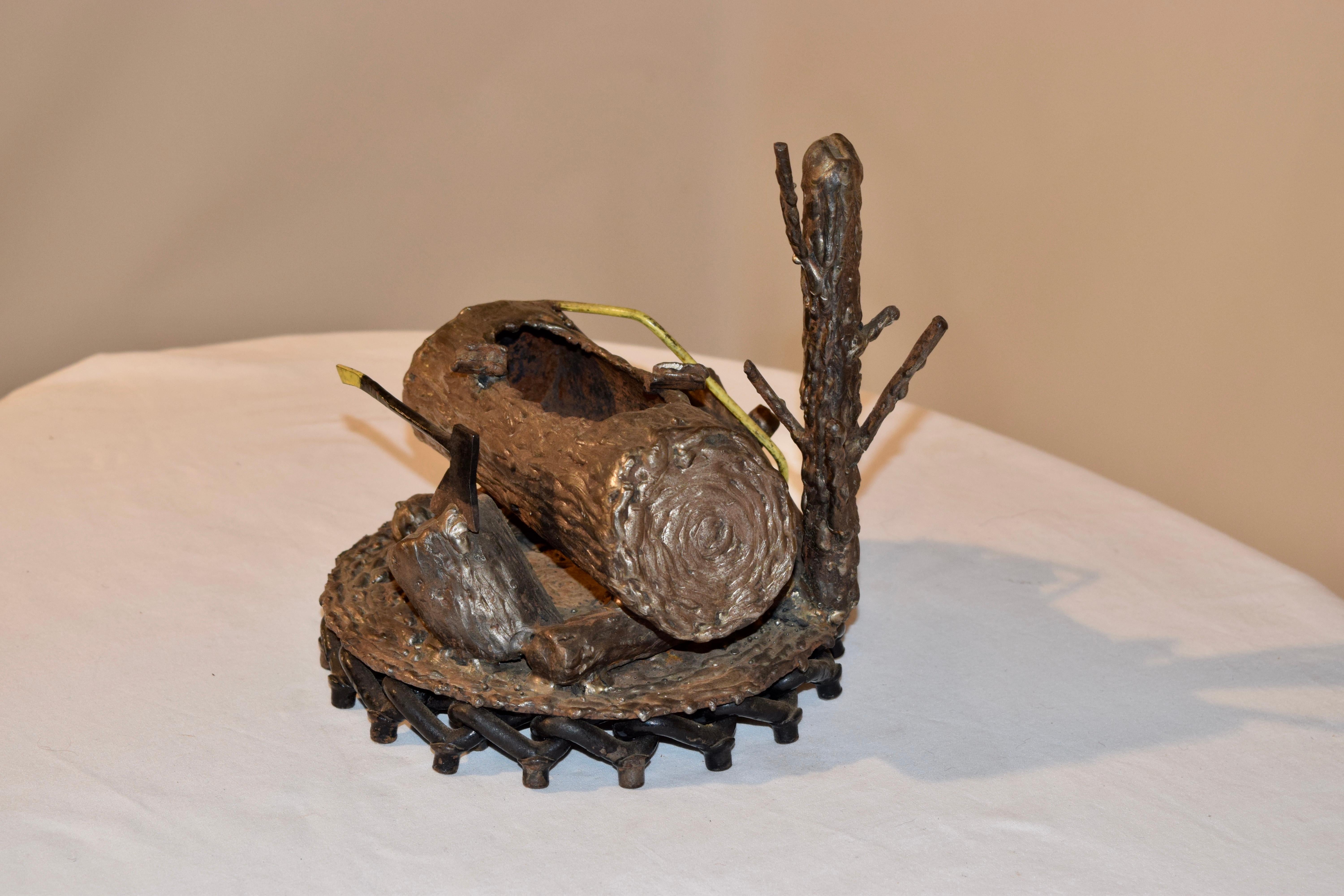 Early 20th century American foundry art ashtray made from iron slag. It is in the form of a tree stump standing behind a large hollowed out log, with a saw resting on one side of the log and an axe stuck in a stump on the other side. It is on an