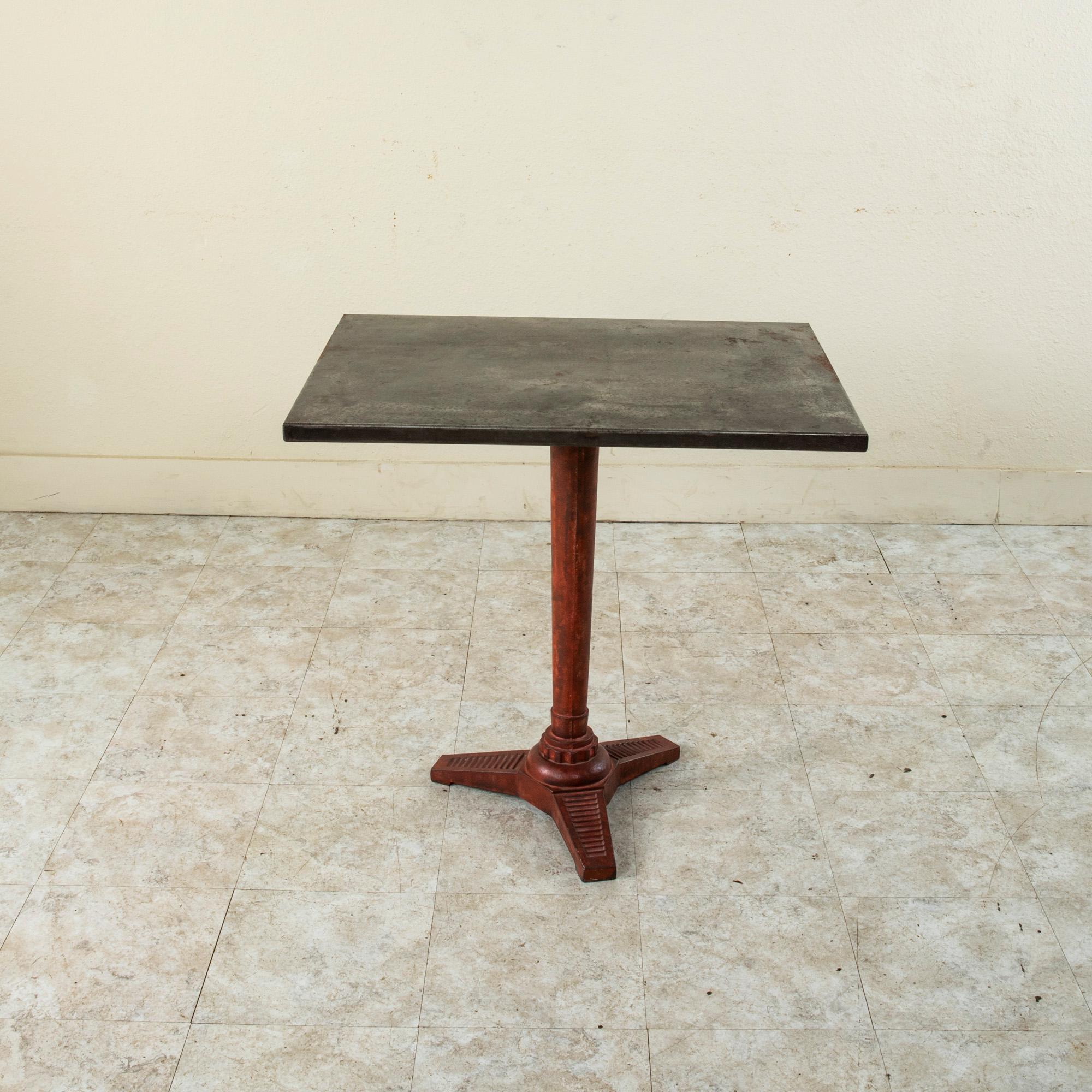 This early twentieth century French Art Deco period rectangular bistro table or cafe table features a metal top on a cast iron pedestal base. The base is painted red and rests on tripod feet detailed with striation. A charming table to lend a bit of