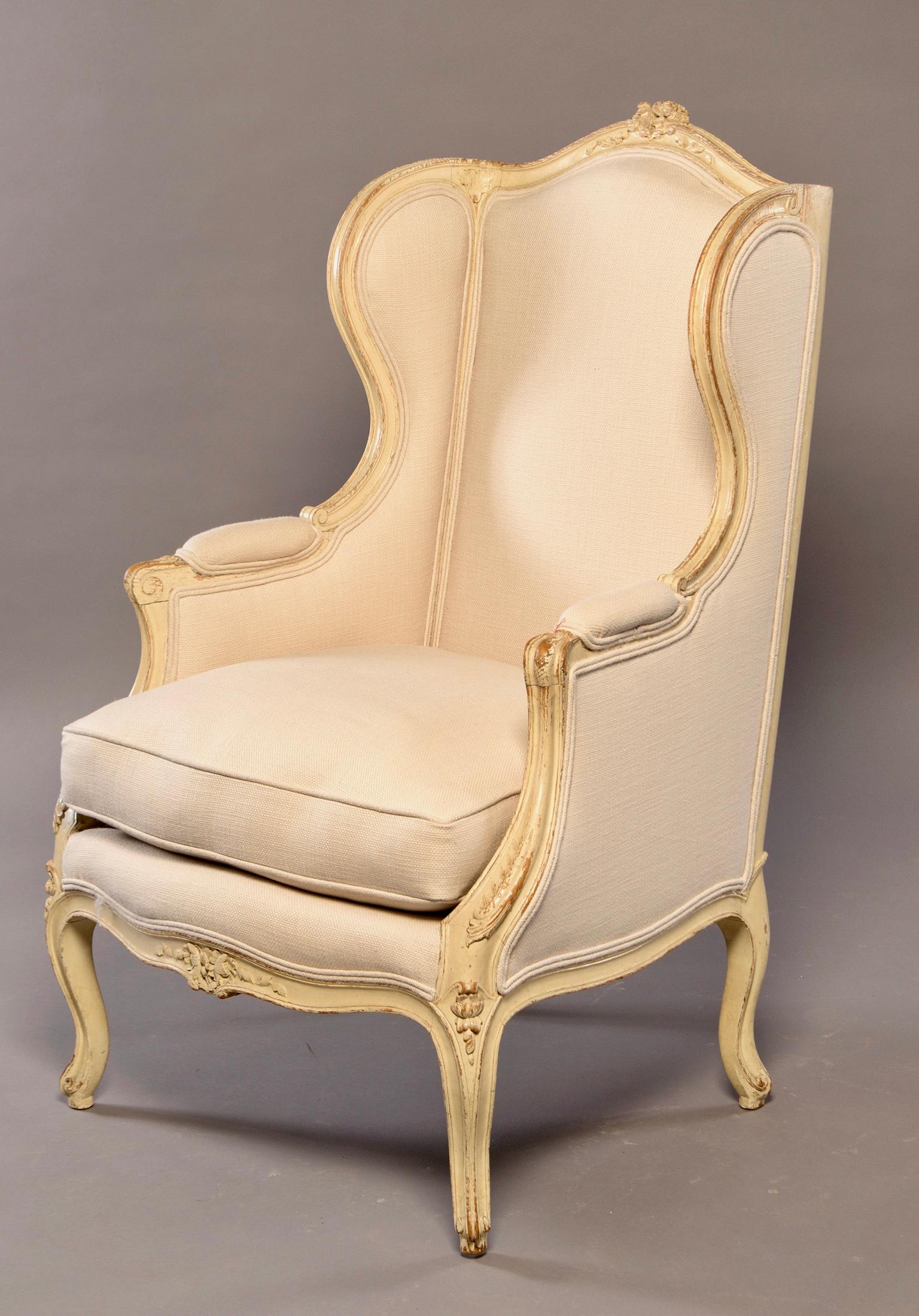 French bergère has carved and painted frame with cabriole legs, and ornamental detail on apron, carved details on the arms and a decorative crest on the seat back, circa 1620. Wing back style with padded arms and newly upholstered in a bone colored