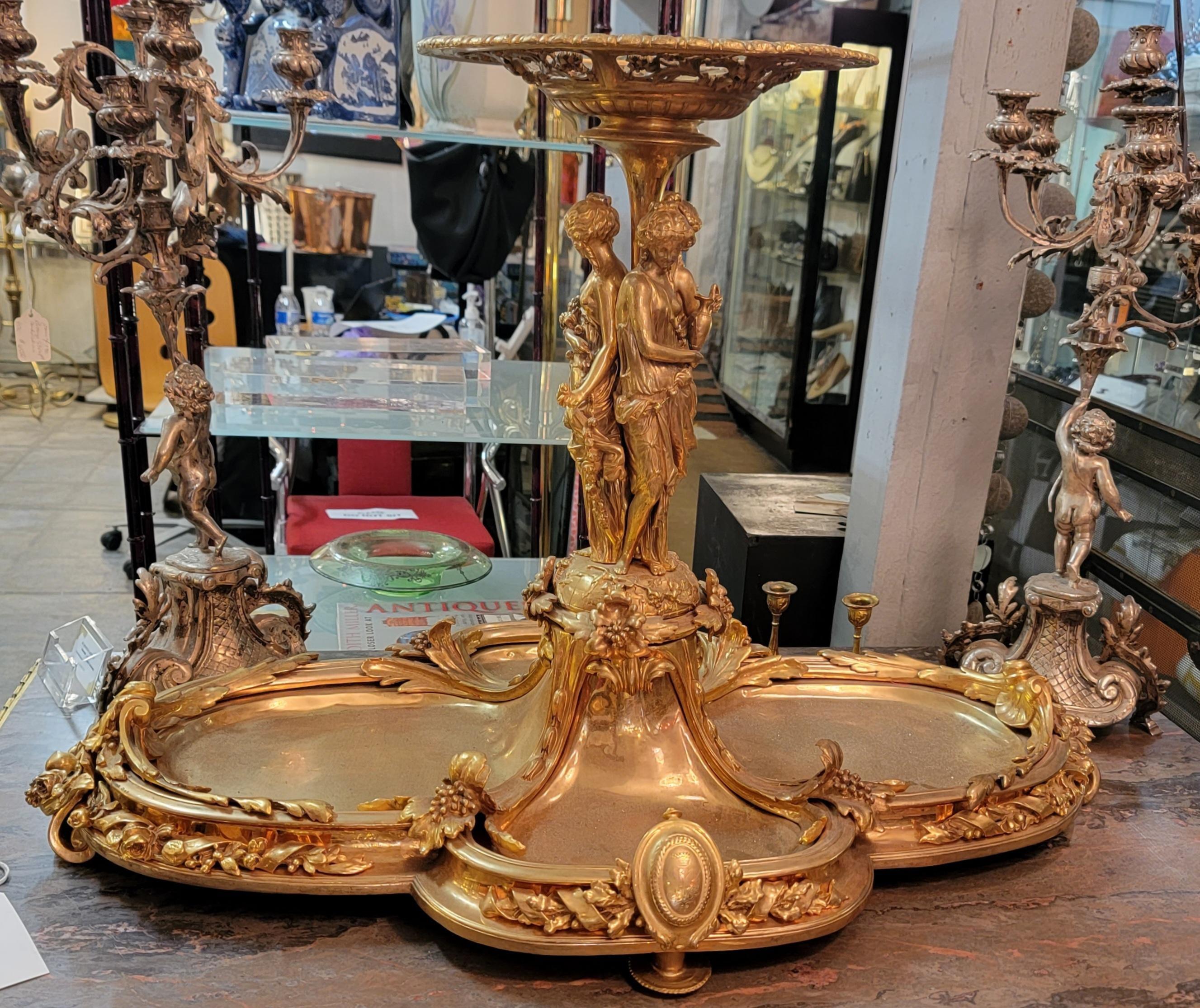 A French Gilt Bronze Figural Centerpiece. Decorated with 3-female figures, each with a basket of flowers or a pitcher. Having a plaque with coat of arms on either side of the plateau.

Dimensions: Height: 24