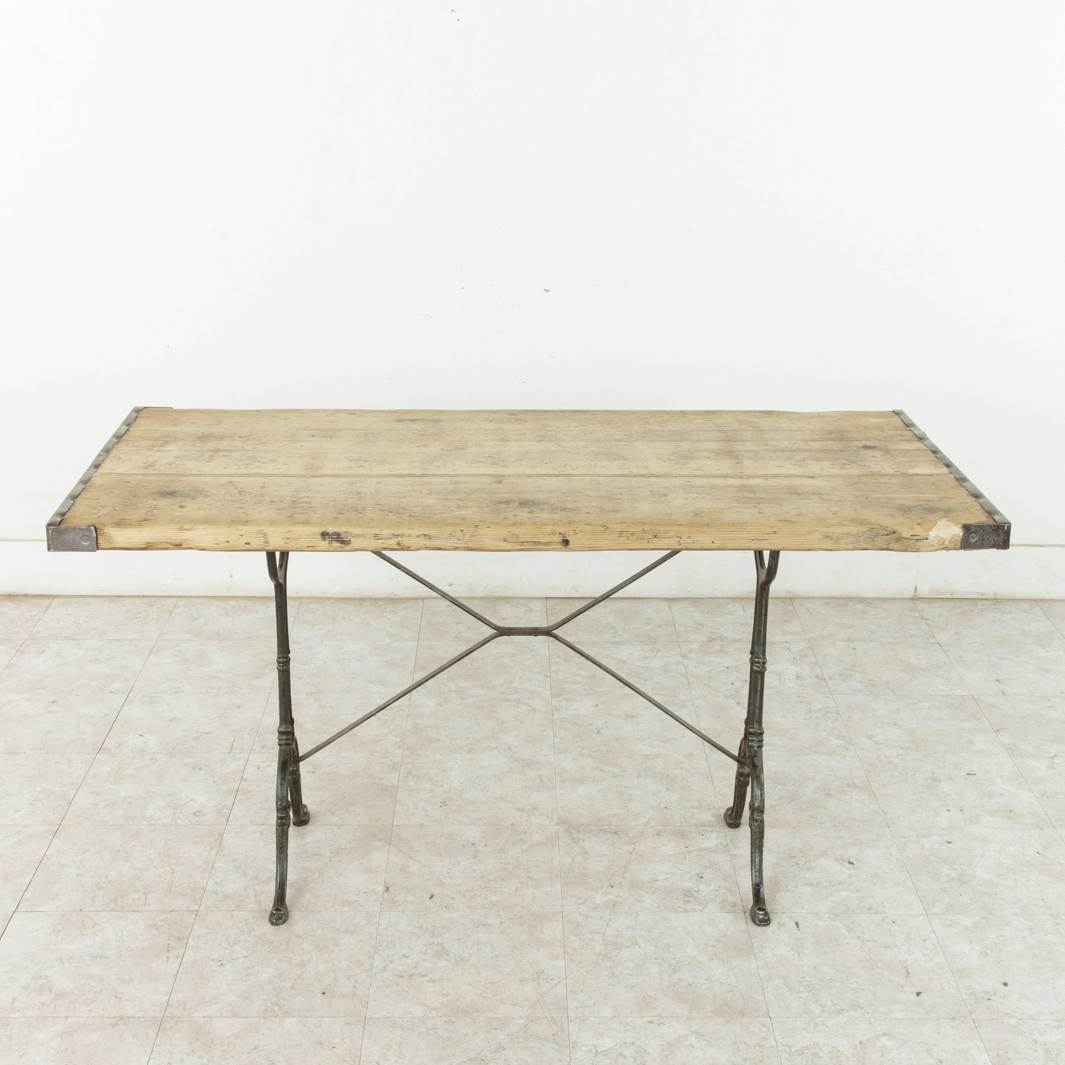 This early 20th century cast iron bistro table features a rustic unfinished pine top trimmed in iron on both sides. The classic scrolled cast iron legs of the piece are joined by an X-stretcher that provides additional stability. Ideal for a