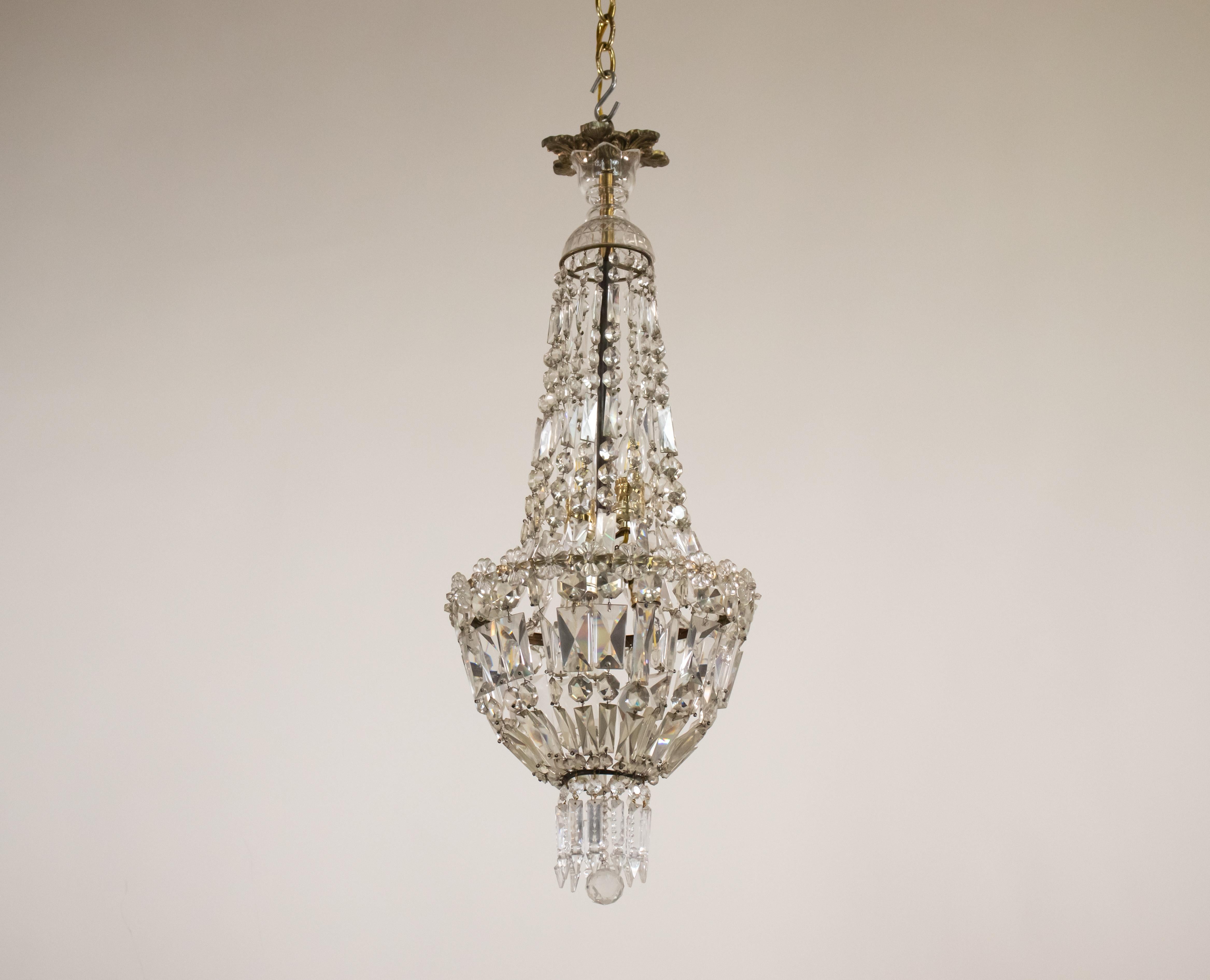 This chandelier features a bronze crown adorned with an acanthus leaf design at its peak. Its structure is beautifully composed of a series of shimmering crystal beads and baguettes that come together to create a crystal bowl, elegantly edged with a