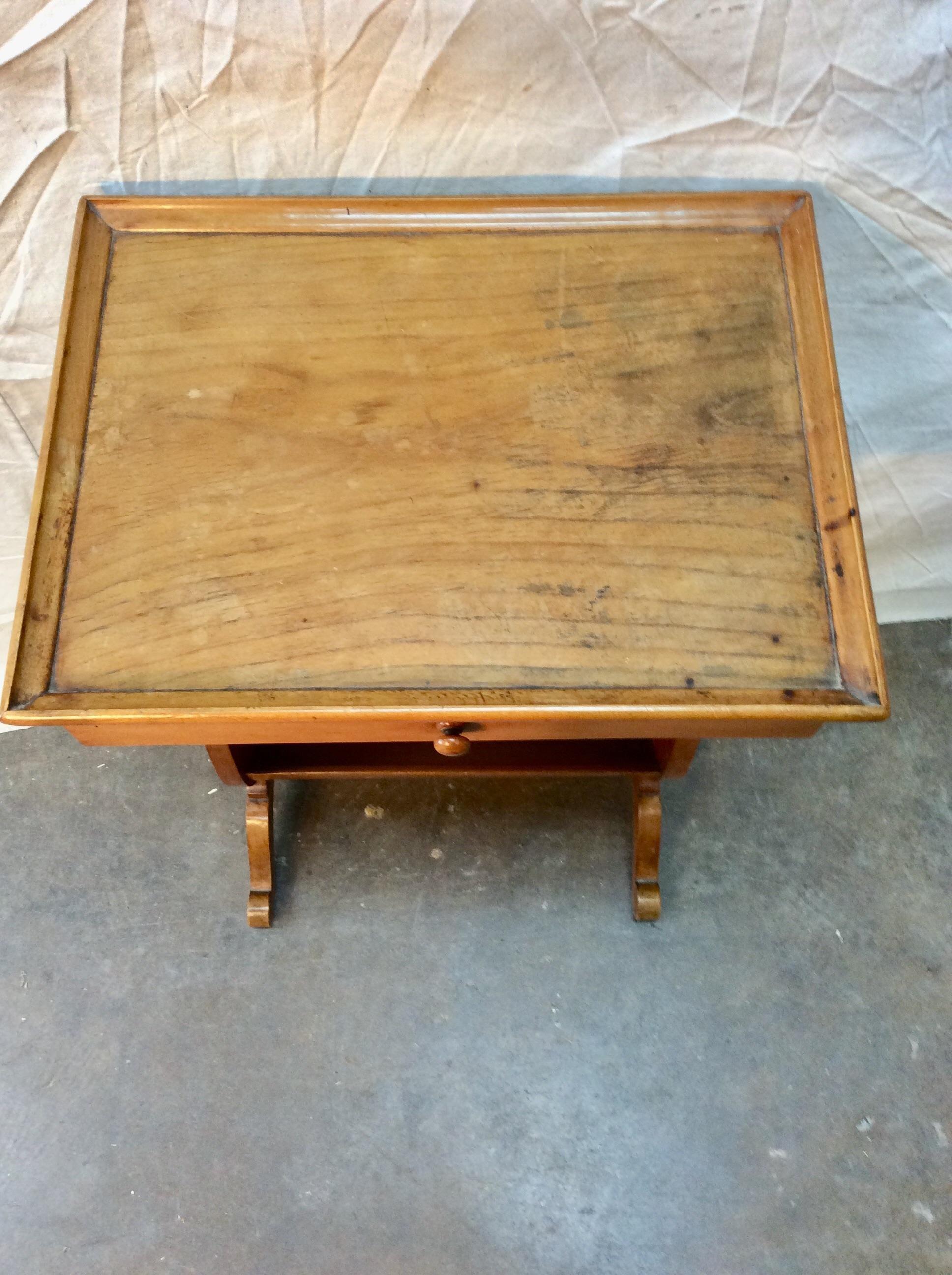 Found in the South of France, this petite early 20th century French Fruitwood Side Table is crafted with a raised edge top, one drawer adorned with a wood pull and a storage space on the bottom, possibly for a book or magazine. The two shapely