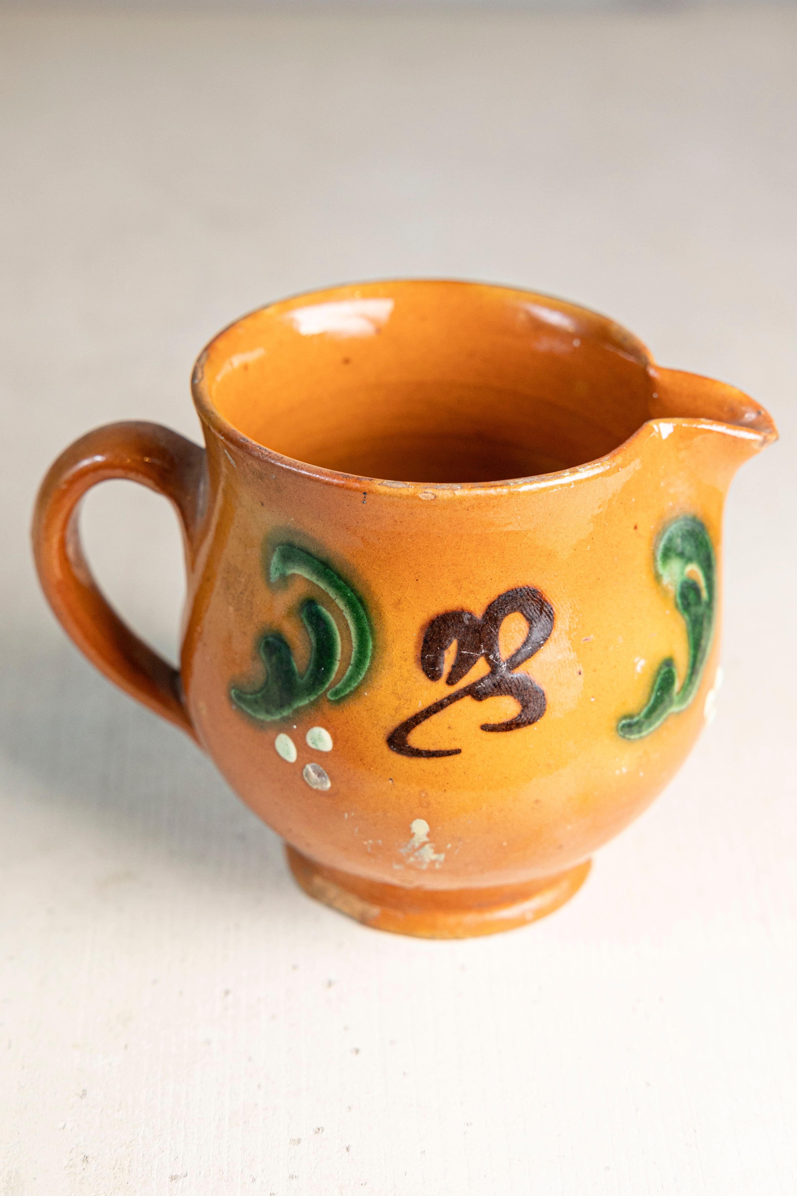 Un petit et charmant pichet en terre cuite française datant du début du 20e siècle, présentant une glaçure ambrée chaude et un décor à l'engobe appliqué à la main en vert, blanc et brun foncé. Mesurant à peine 4,5 pouces de large, ce petit récipient