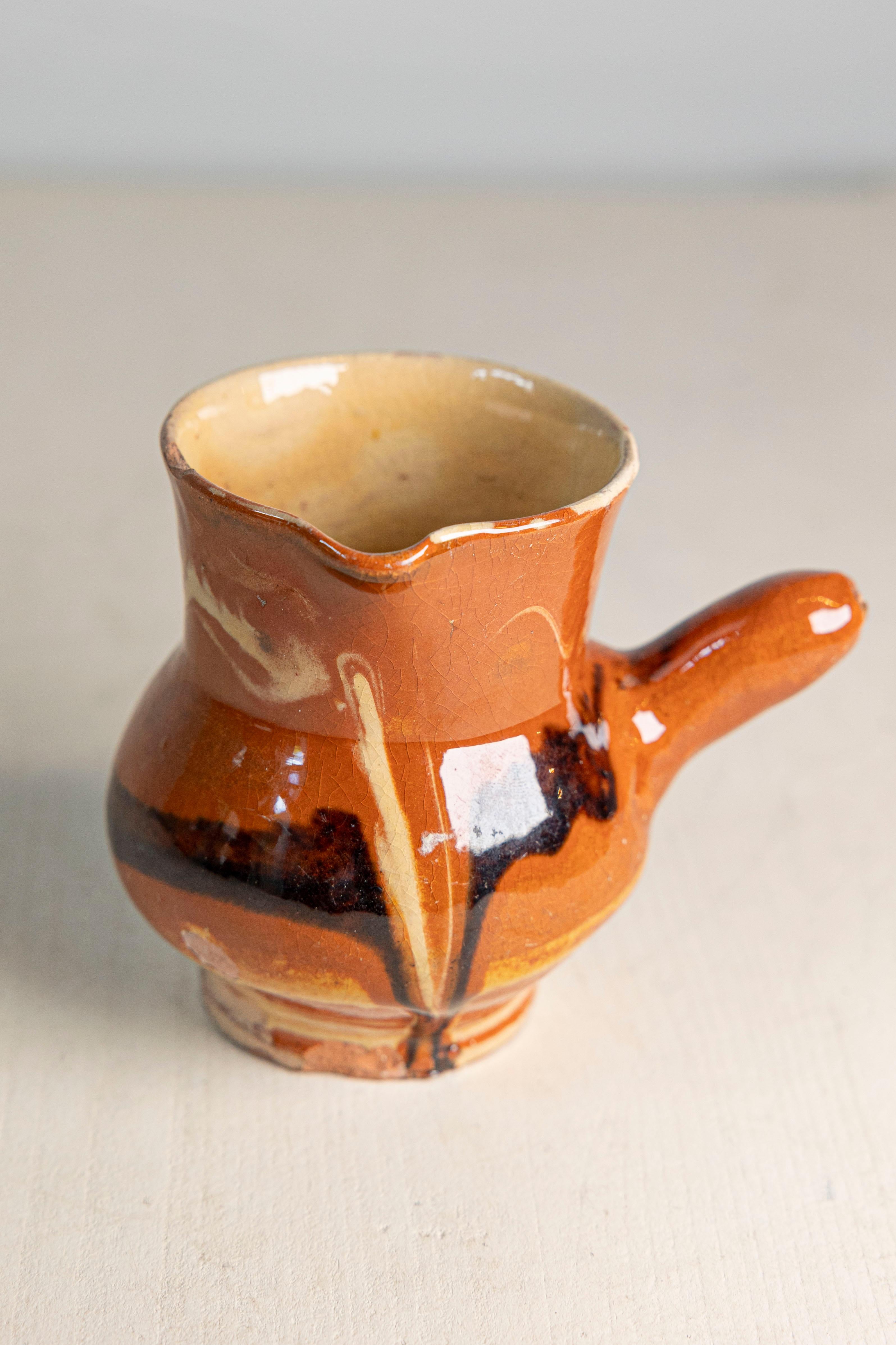 Charmant pichet rustique en terre cuite française du début du 20e siècle, tourné à la main et fini selon la technique traditionnelle de l'engobe. La pièce présente un petit corps arrondi, un bord légèrement évasé avec un bec verseur et une poignée