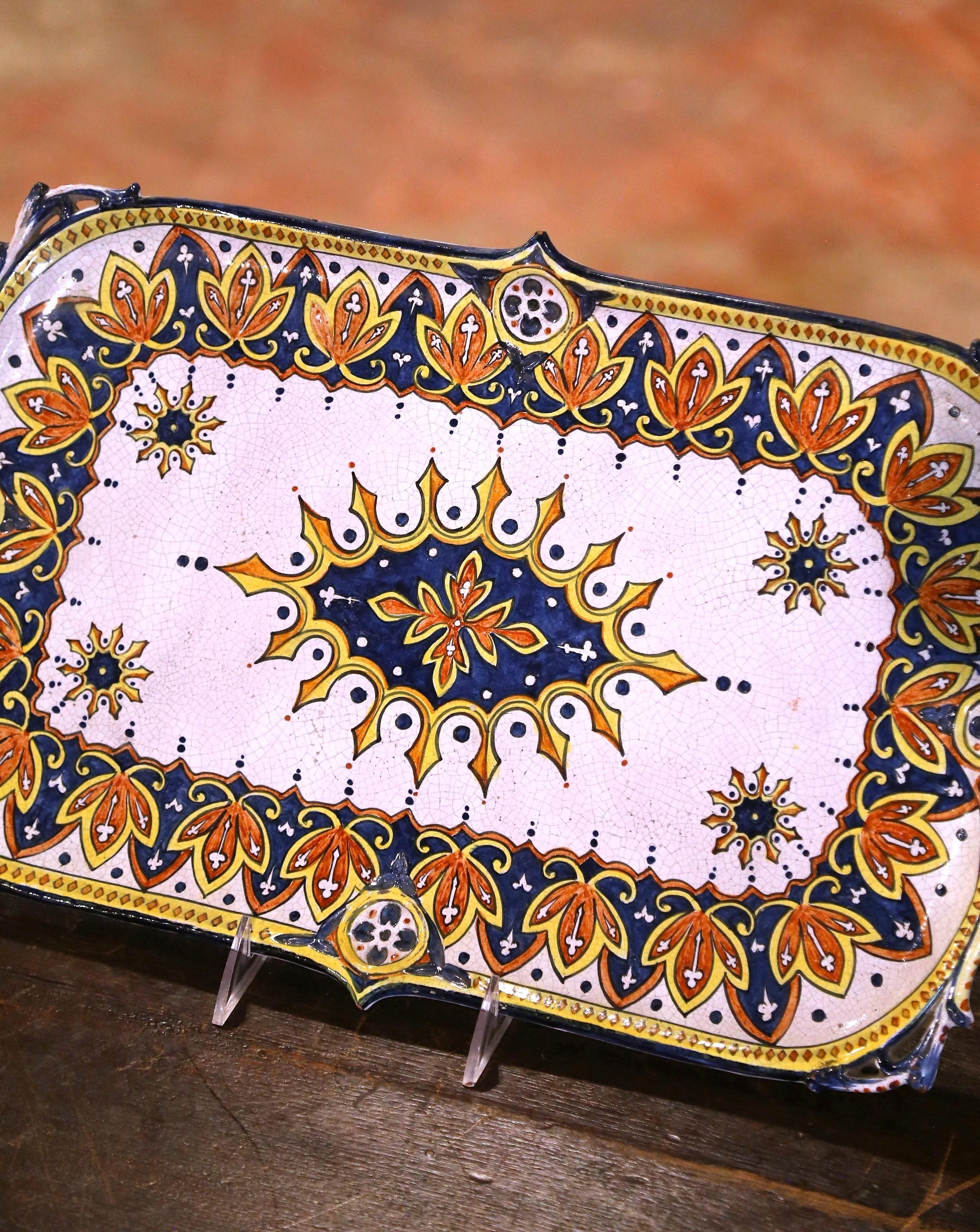 Sirve a tus invitados con estilo con esta exquisita fuente de loza francesa antigua. Fabricada en Rouen, Francia, hacia 1930, esta obra maestra de cerámica pintada a mano presenta un llamativo medallón central con motivos radiantes, enmarcado por un