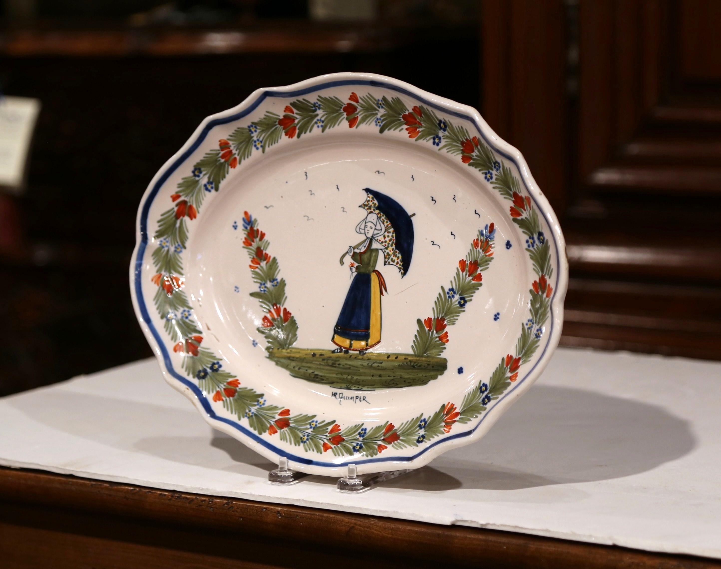 This colorful ceramic wall plate was created in Brittany, France, circa 1904-1922. Oval in shape, the faience platter features hand painted decor including a traditional Breton woman figure dressed in authentic costume and holding an umbrella. The