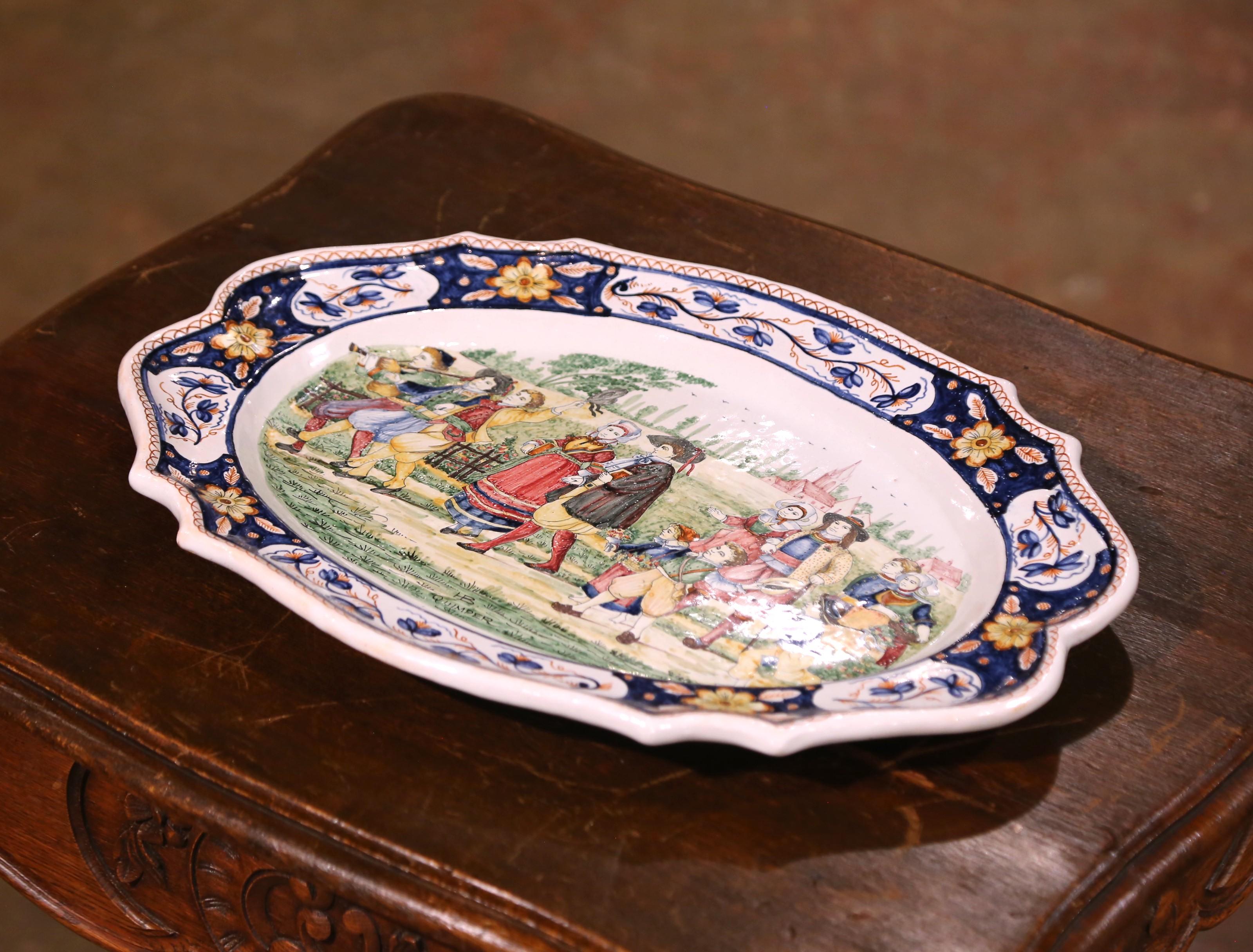 Ce plat en céramique antique a été fabriqué en Bretagne, en France, vers 1920, et illustre l'esprit vivant de la faïence de Quimper. De forme ovale avec un bord festonné, cette pièce en terre cuite vernissée à l'étain est peinte à la main d'une