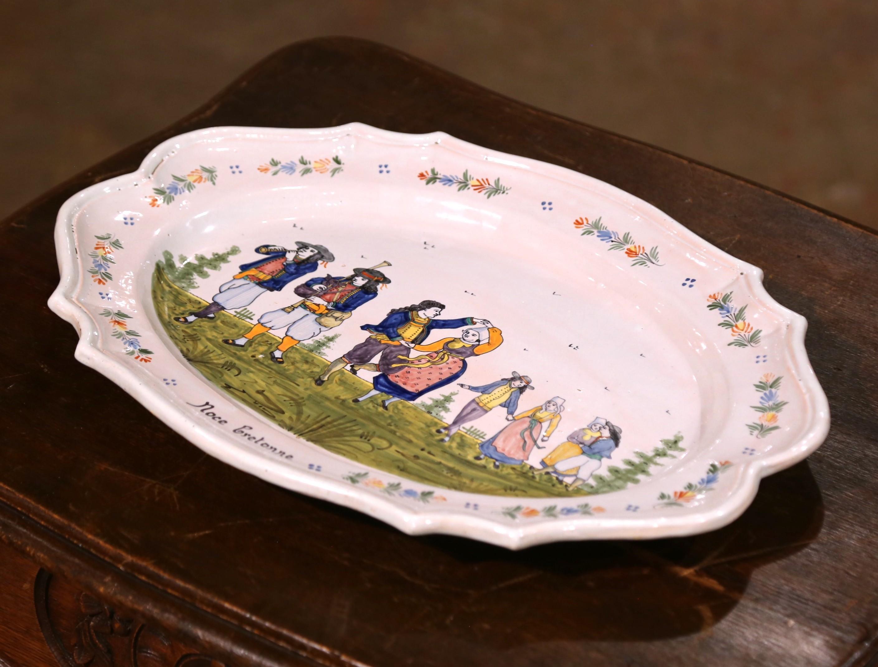 This charming antique wall platter was hand-painted in Brittany, France, circa 1900 by the HB Quimper workshop. Oval and scalloped in form, the tin-glazed earthenware piece features a lively and colorful scene titled 