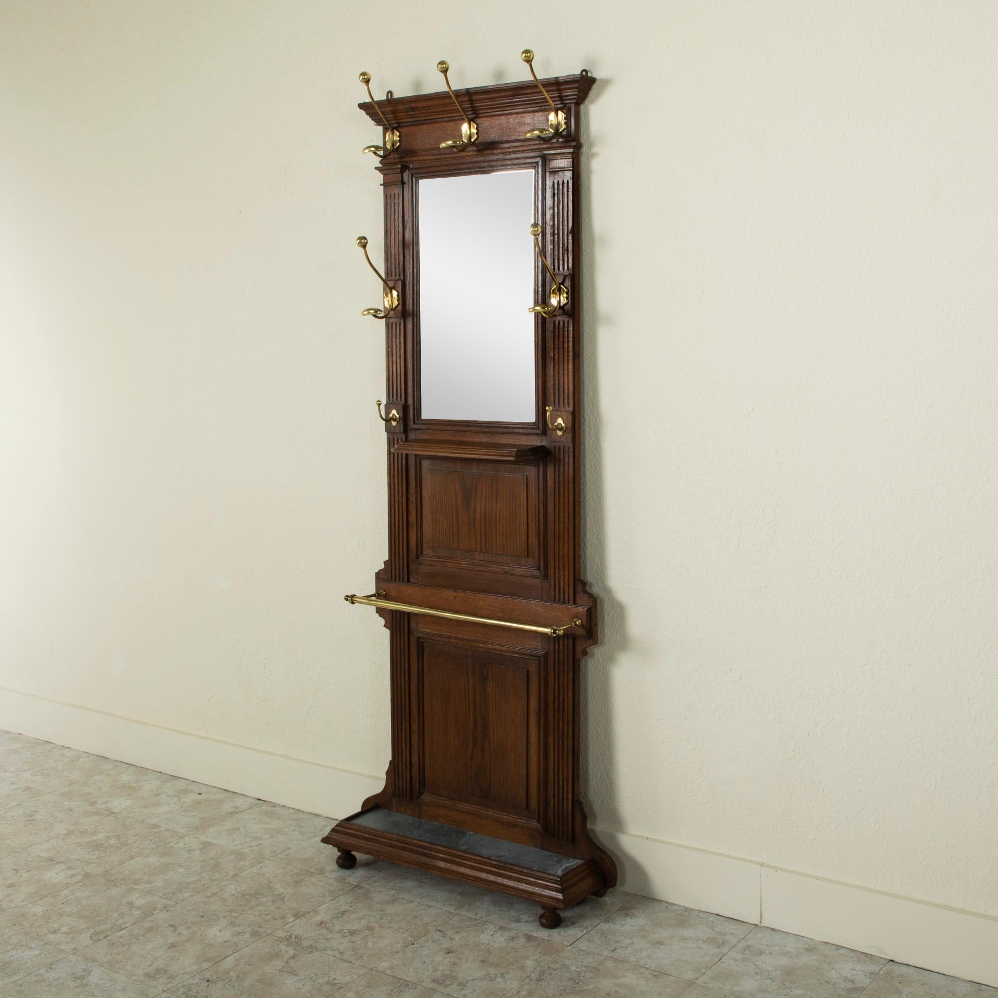 This handsome late nineteenth century Henri II style hall tree is constructed of solid oak with seven bronze hooks and its original beveled mirror. Of a versatile, narrow scale, this piece features an upper row of three hat hooks and two additional