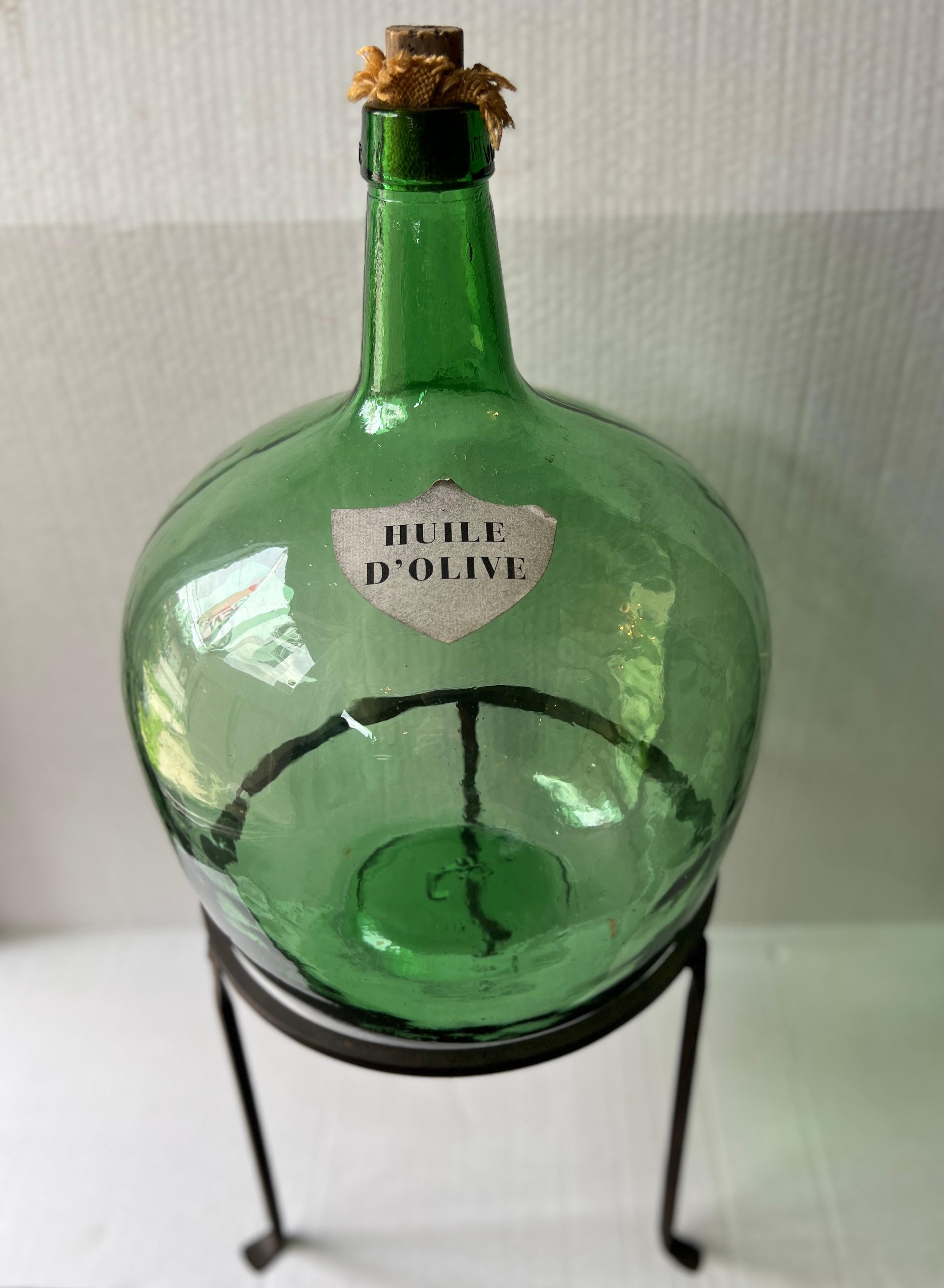 French Huile D'olive Green Glass Demijohns are charming, vintage glass containers that once held aromatic olive oil, now serving as elegant décor pieces.

The wrought iron stand is included.