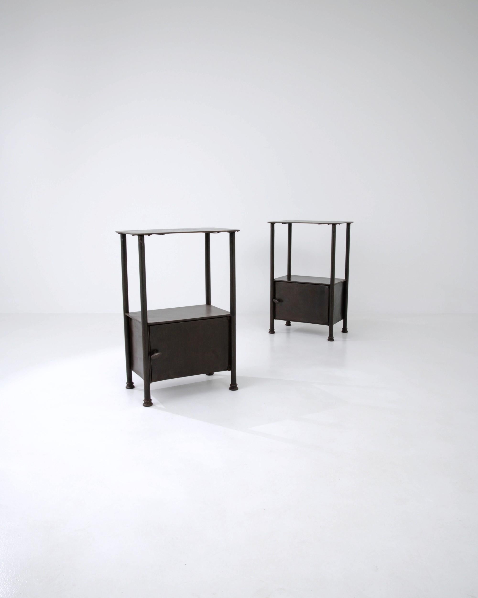 Combining a bold form with a dark patina, this pair of vintage metal bedside tables make an attractive Industrial accent. Built in France in the early 20th century, a low cupboard is framed by the tall pillars of the legs, which rise to support an