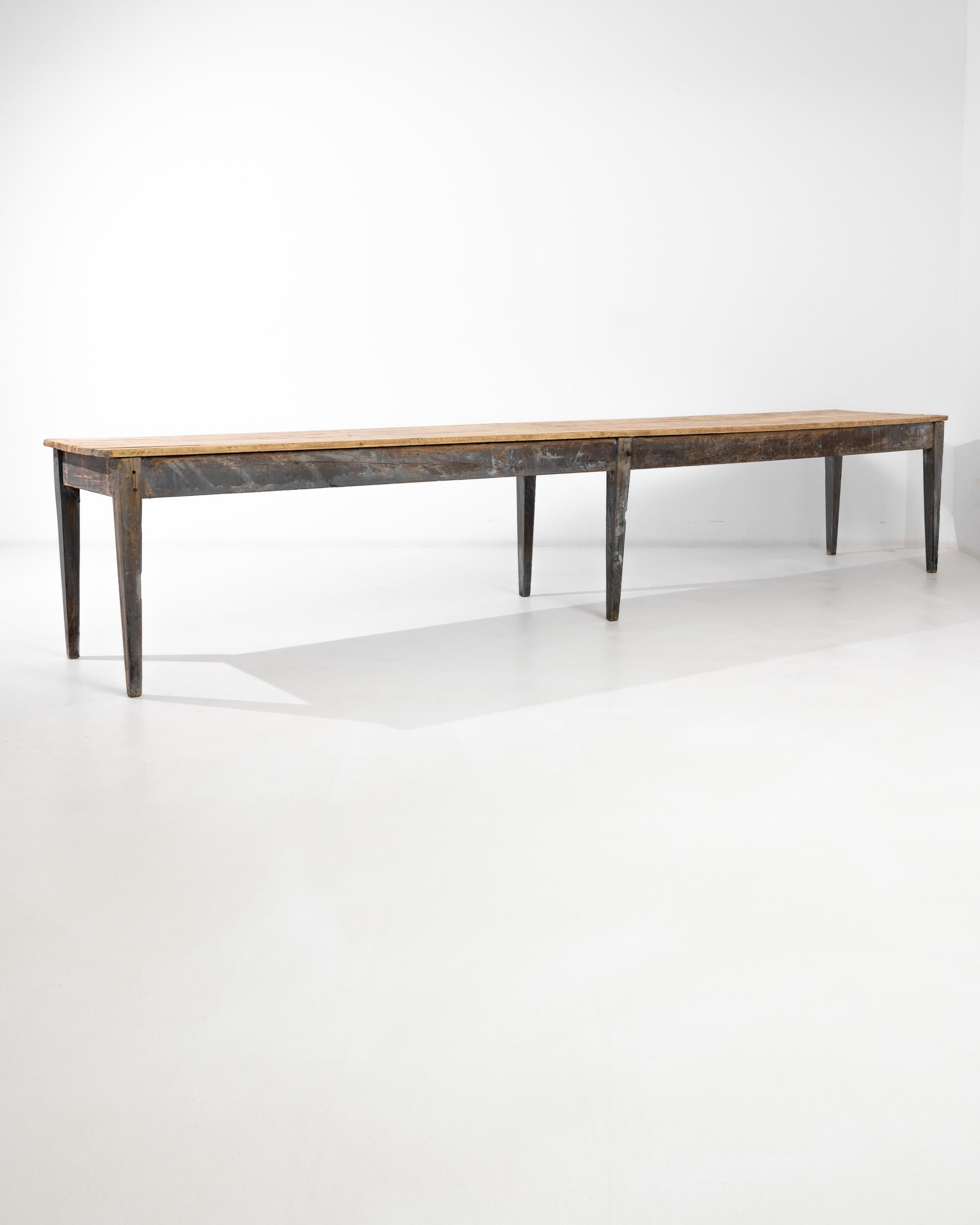 This impressively long wooden table was made in France circa 1900. Three pairs of legs with tapered feet are attached to a stout and wide apron. The blonde rustic tabletop lends a refreshing counterpoint, contrasting with the patinated gray finish