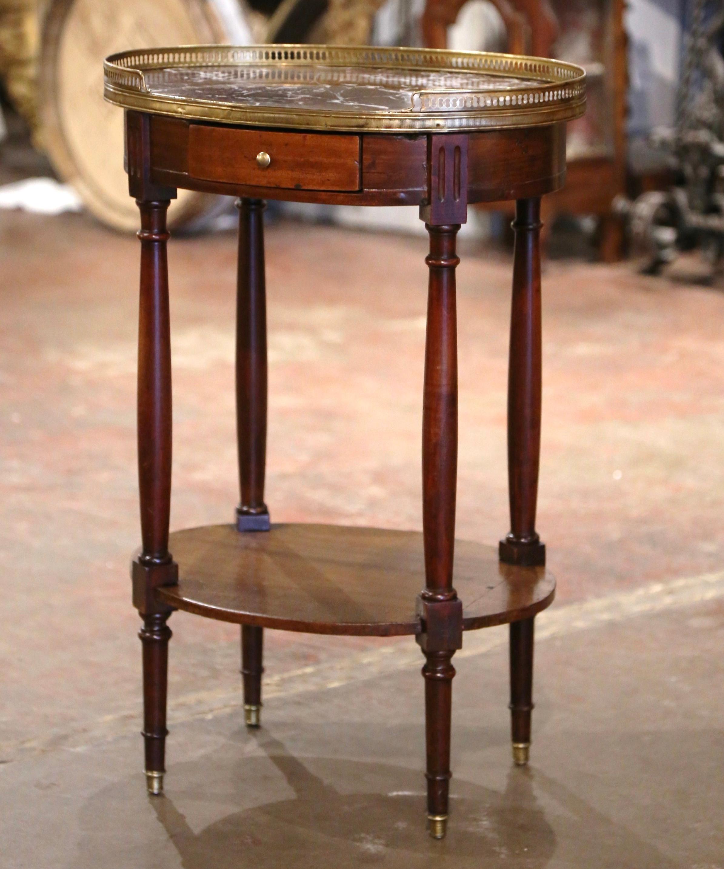 Crafted in France circa 1920 and built of walnut, the oval table sits on four turned legs decorated with brass mounts at the feet over a curved apron dressed with a drawer. The luxurious variegated brown and white marble top is set inside an