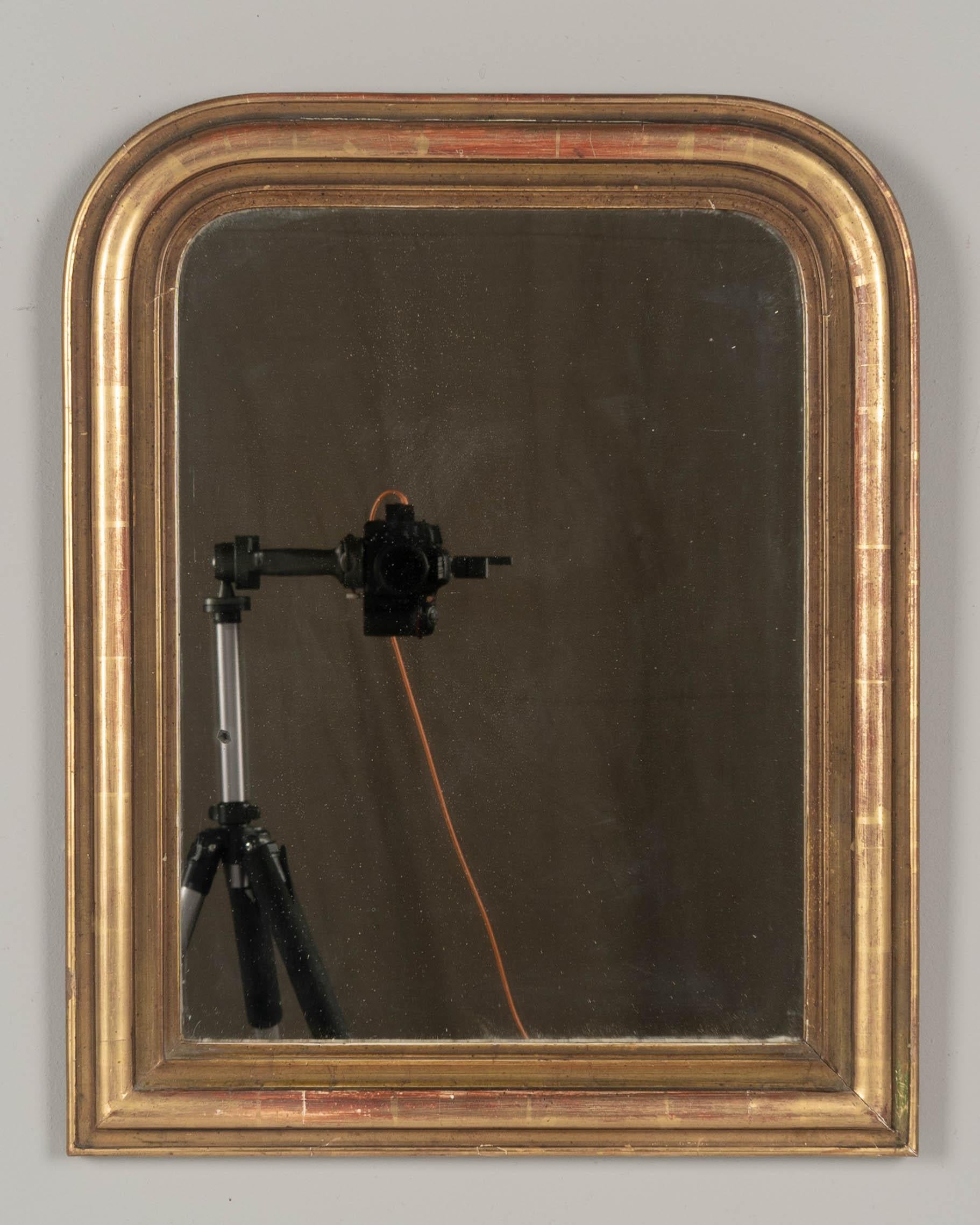 An early 20th century French Louis Philippe style small gilded mirror with curved top corners. Warm gilt finish with red under rub exposed. Original mirror with some spotting. Please refer to photos for more details. Pictures are part of the