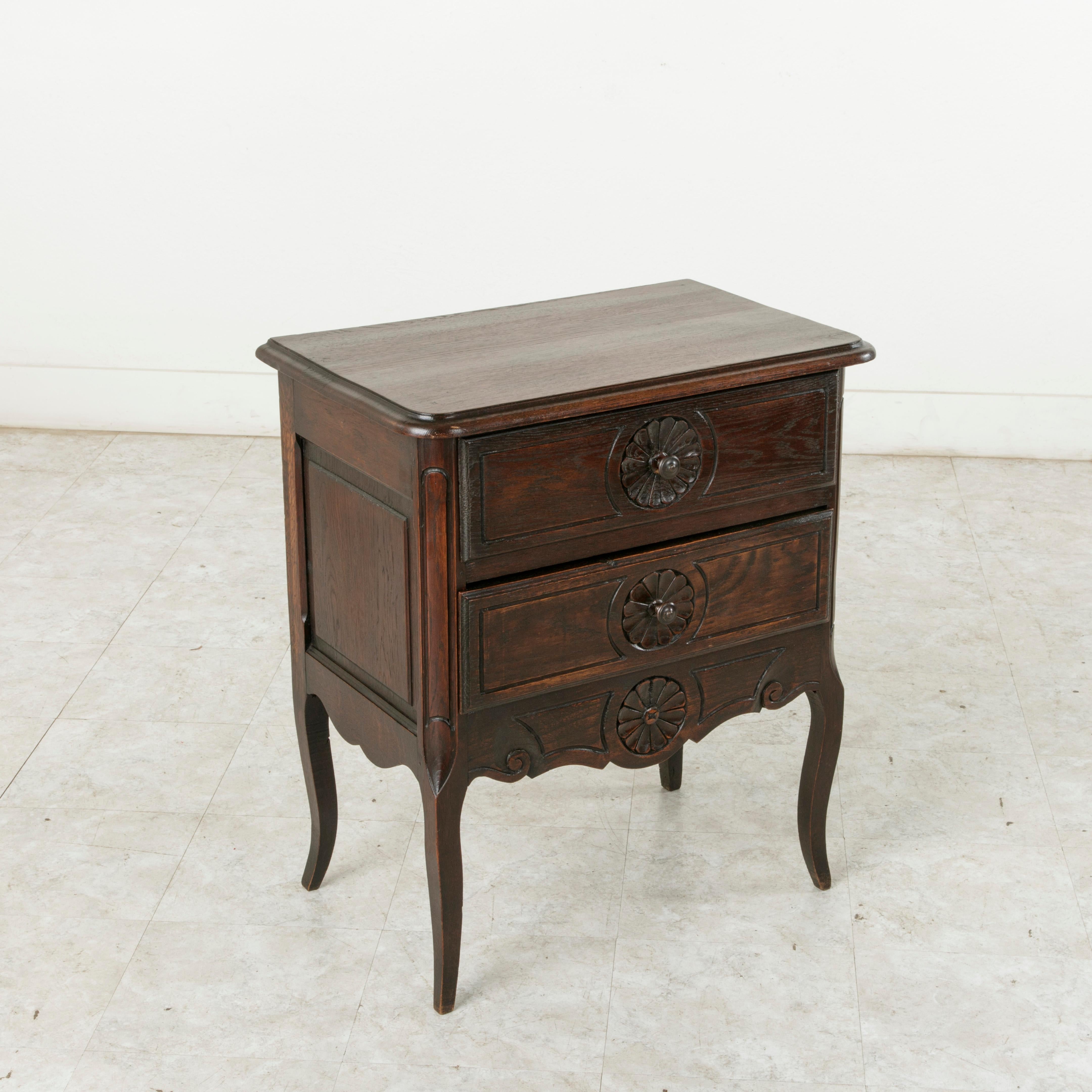 This early 20th century French Louis XV style small-scale oak commode or chest features hand-carved rosettes on its two drawer fronts around the iron drawer pulls and a carved rosette in the lower apron. The beveled top and paneled sides of the