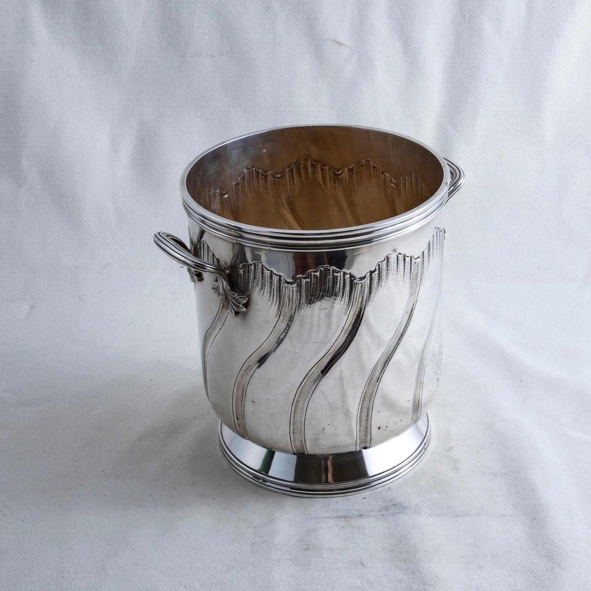 This early twentieth century French silver plate champagne bucket features a classic Louis XV style spiraling scallop on the sides. The bucket is appointed with a handle on each side detailed with leaves and rests on a footed base. The bottom of the