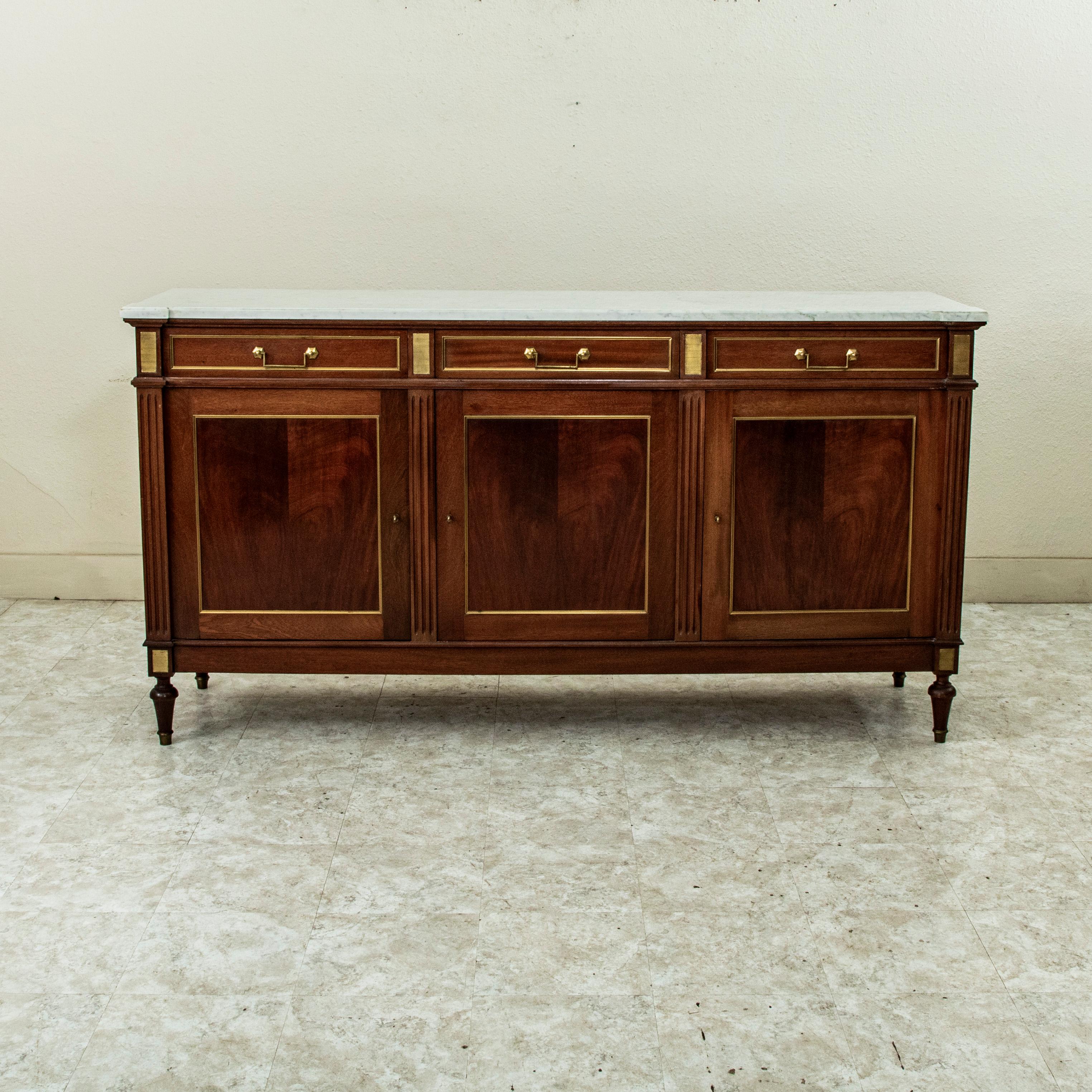 This early twentieth century Louis XVI enfilade, or sideboard is constructed of solid mahogany and features a beveled white marble top with grey veining. This piece has bronze trim that surrounds each panel on the sides and front and classic Louis