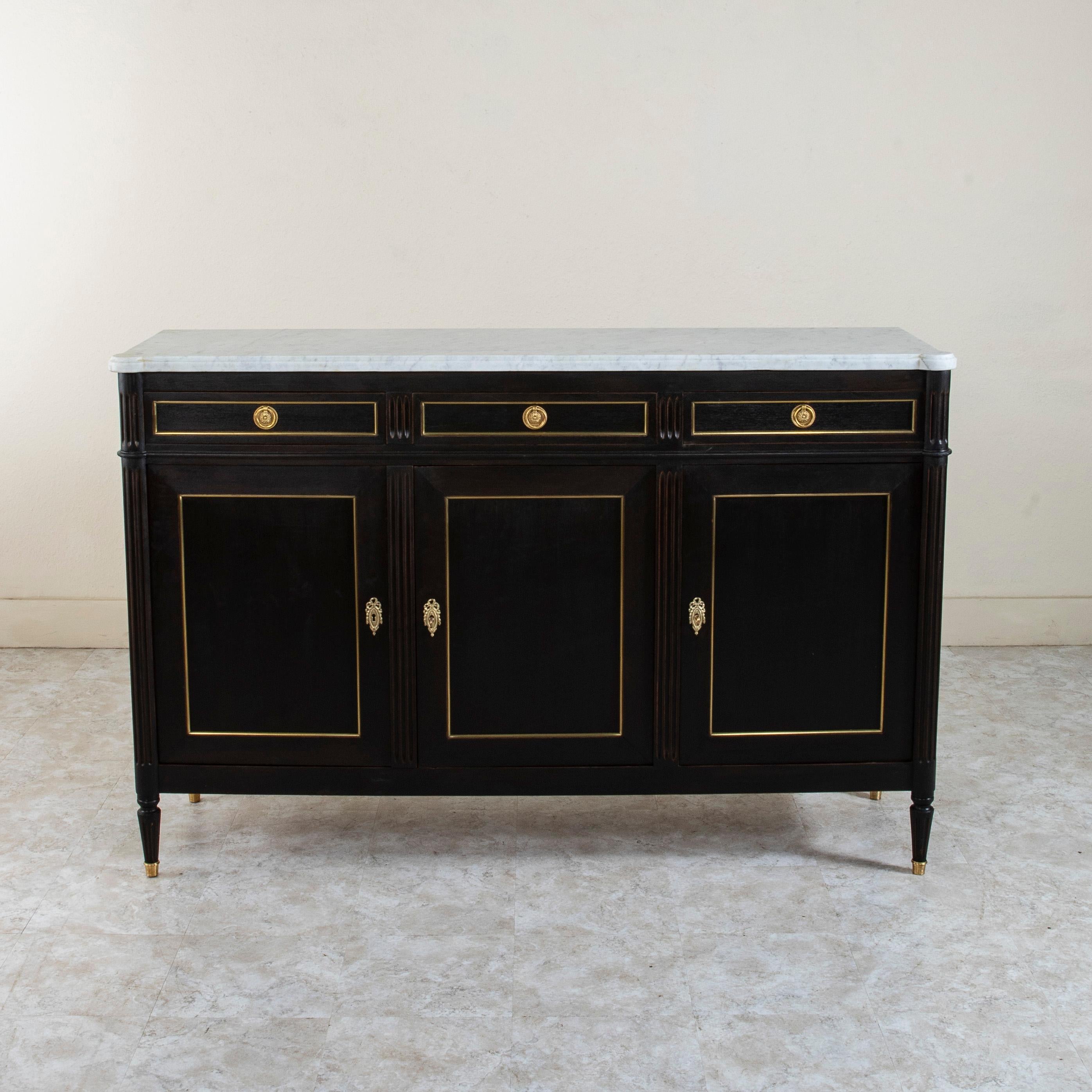 This early twentieth century French Louis XVI style enfilade or sideboard features a black painted mahogany cabinet with a white beveled marble top. Stunning bronze banding outlines each door and drawer, while fluted columns define the facade. Its