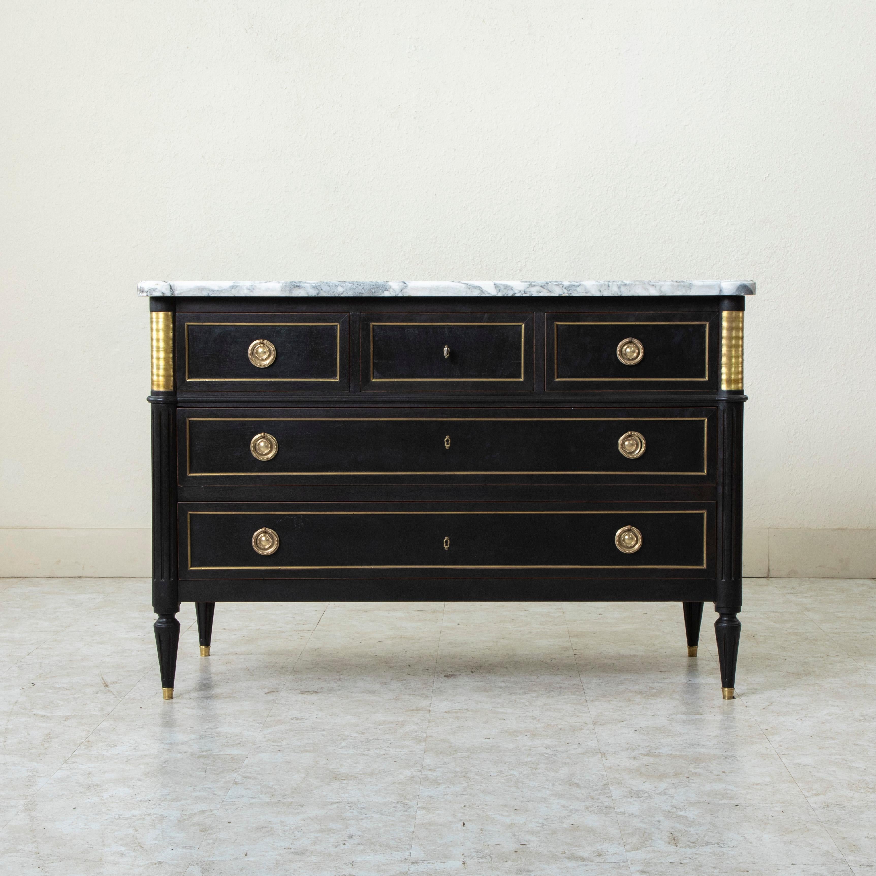 This early 20th century French Louis XVI style black painted ash commode or chest features a beveled white marble top with grey veining. The rounded front corners are detailed with fluting and striated bronze plaques. Each panel is surrounded by a