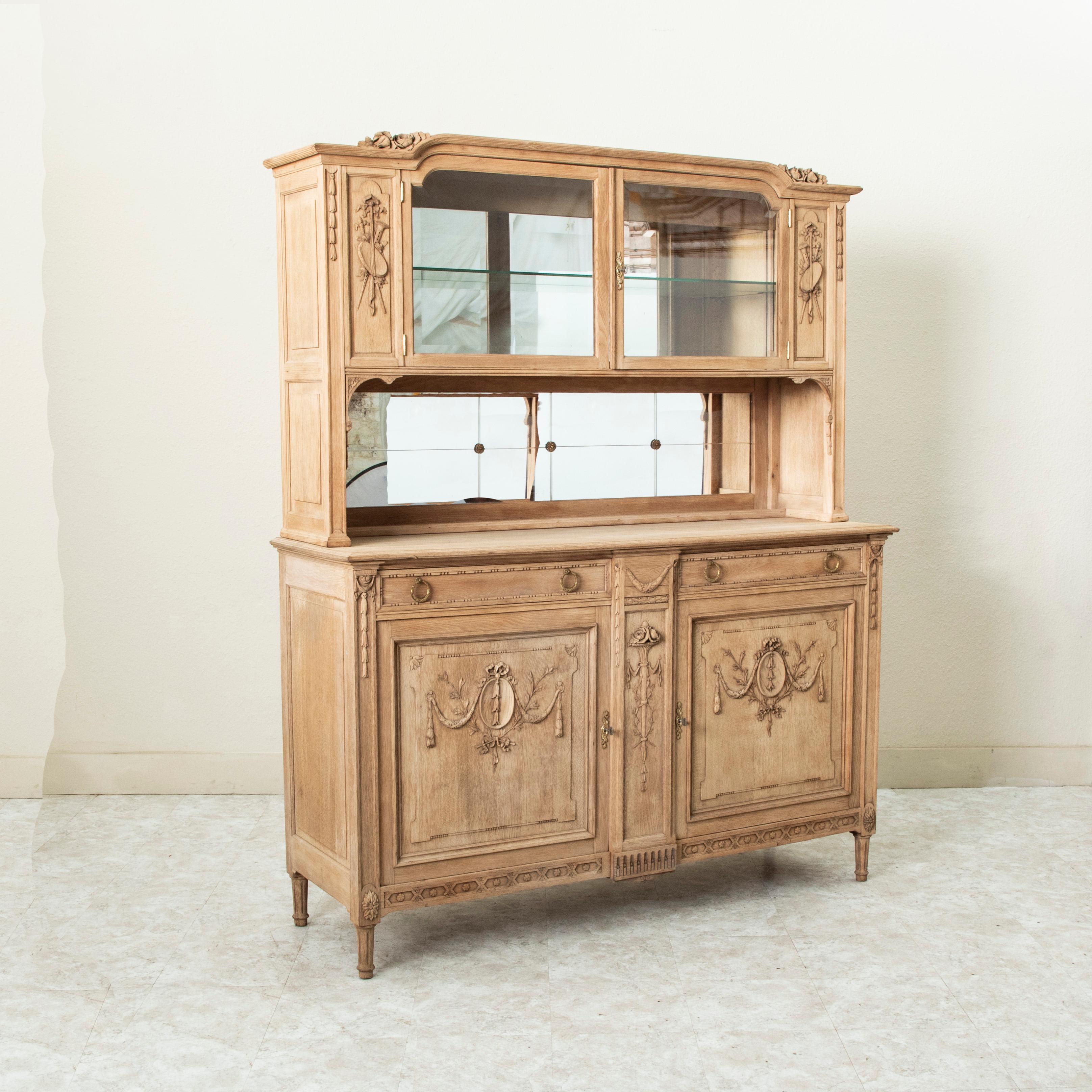Este buffet deux corps francés de principios del siglo XX, de estilo Luis XVI, presenta detalles tallados a mano de instrumentos musicales, guirnaldas de guirnaldas y un motivo floral. El armario superior está equipado con dos puertas de cristal que