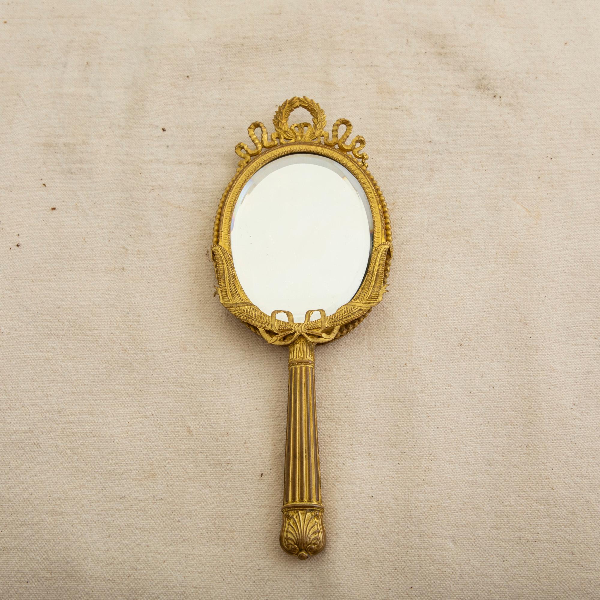 This early twentieth century French Louis XVI style gilt bronze hand mirror features a laurel wreath at the crown tied with a scrolling knotted ribbon. Feathers flank the bottom edge and are additionally tied with a classic knotted ribbon.