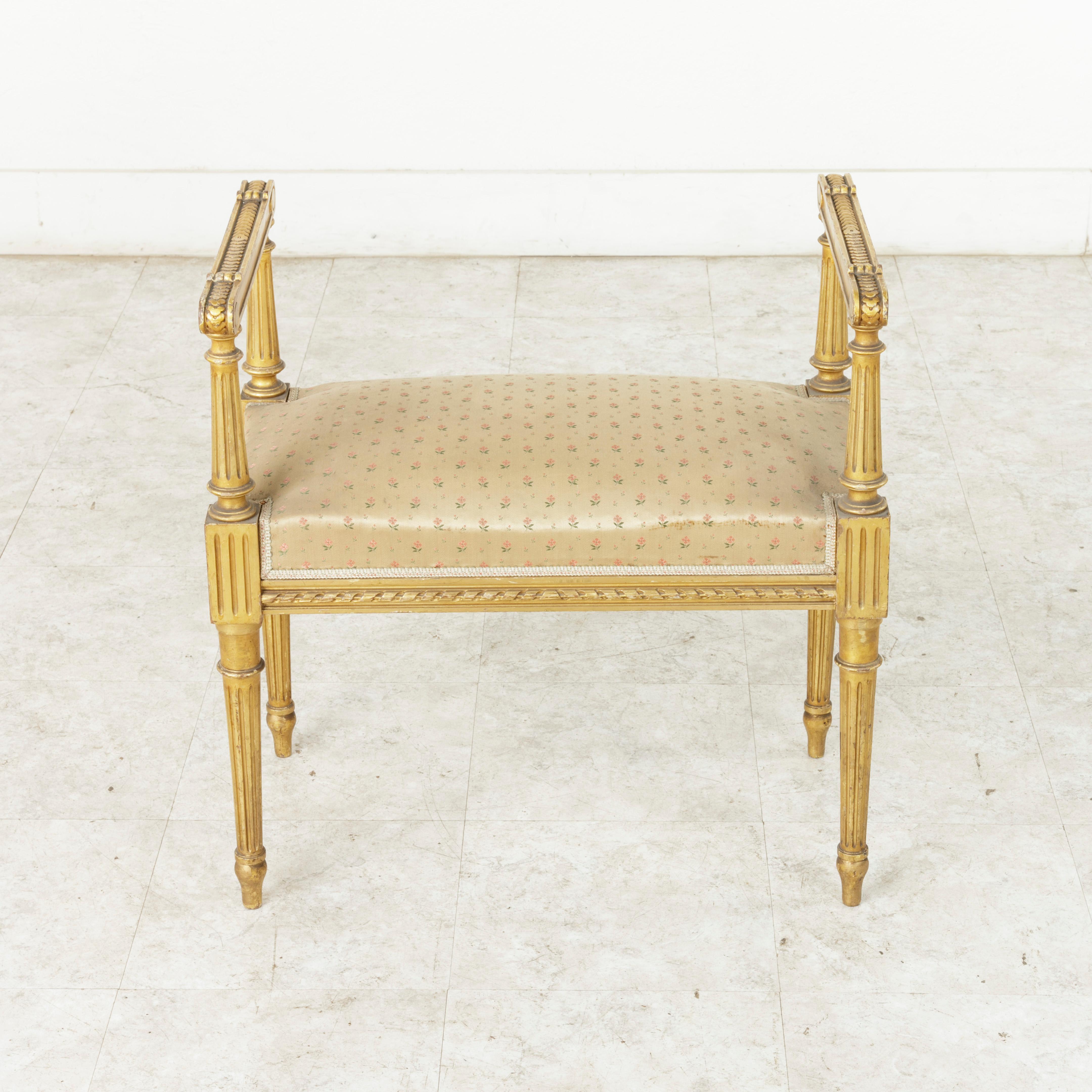 This French Louis XVI style giltwood vanity stool from the early 20th century features carved overlapping quatrefoils on the scrolled armrests and twisted ribbon around the apron. Its tapered fluted legs support the fluted die joints, and fluted