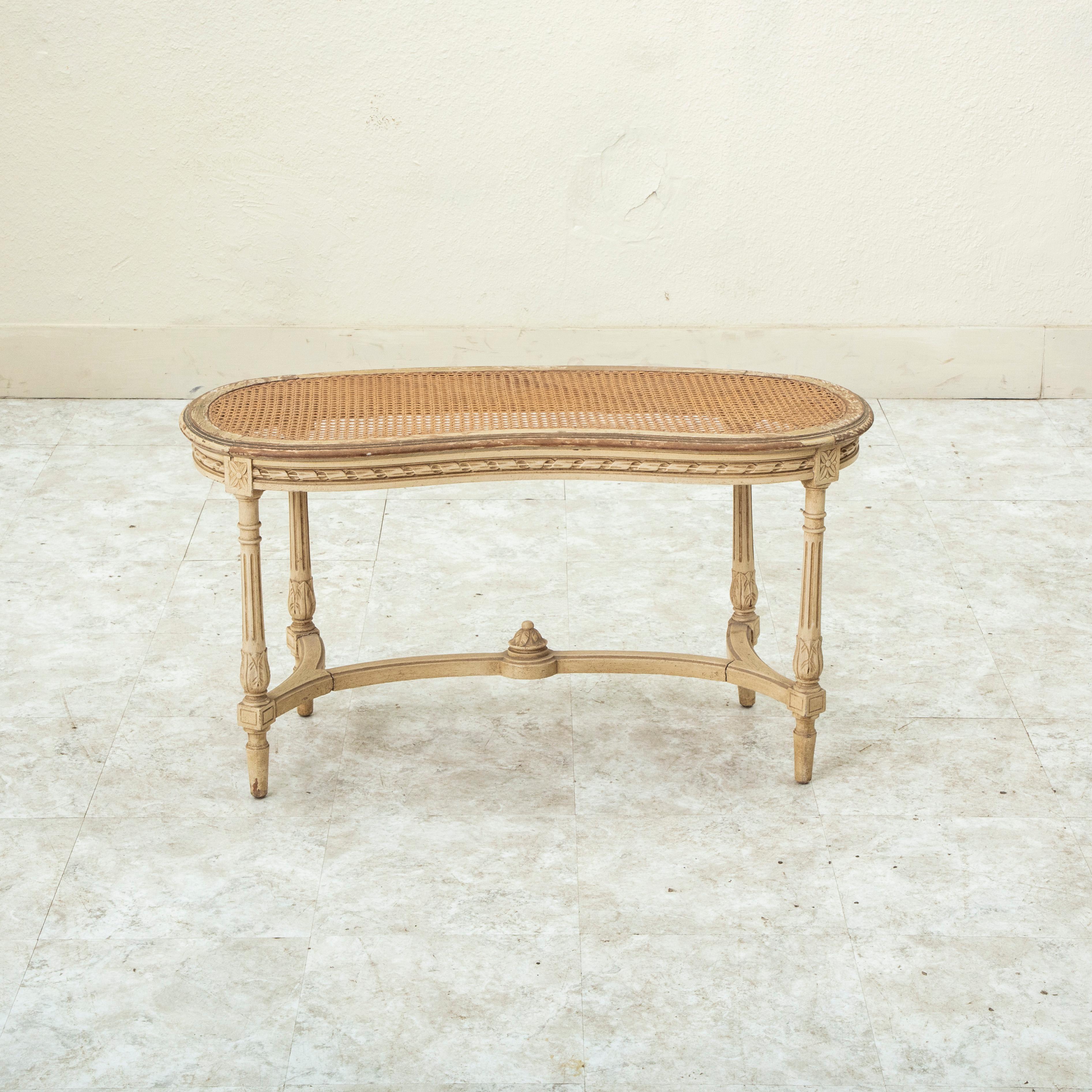 Cette banquette en hêtre peint de style Louis XVI, datant du début du XXe siècle, présente des motifs classiques sculptés à la main, tels que des rosettes au niveau des joints et un motif de ruban torsadé le long du tablier. Des feuilles d'acanthe