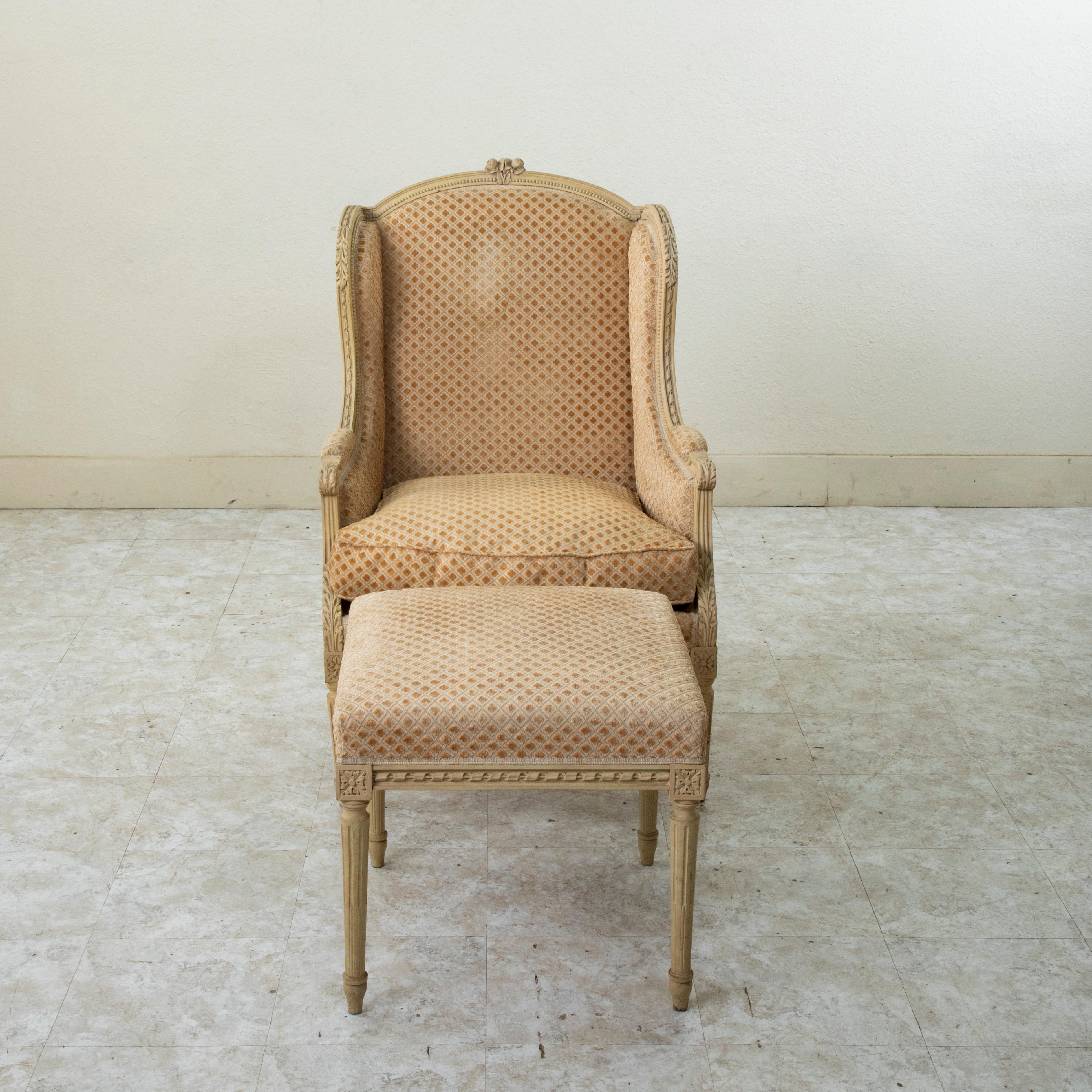 Ce fauteuil ou bergère à oreilles peint de style Louis XVI français du début du vingtième siècle comprend son ottoman. Le fauteuil présente des détails sculptés à la main, tels que des fleurs au niveau de la couronne et des feuilles d'acanthe sur
