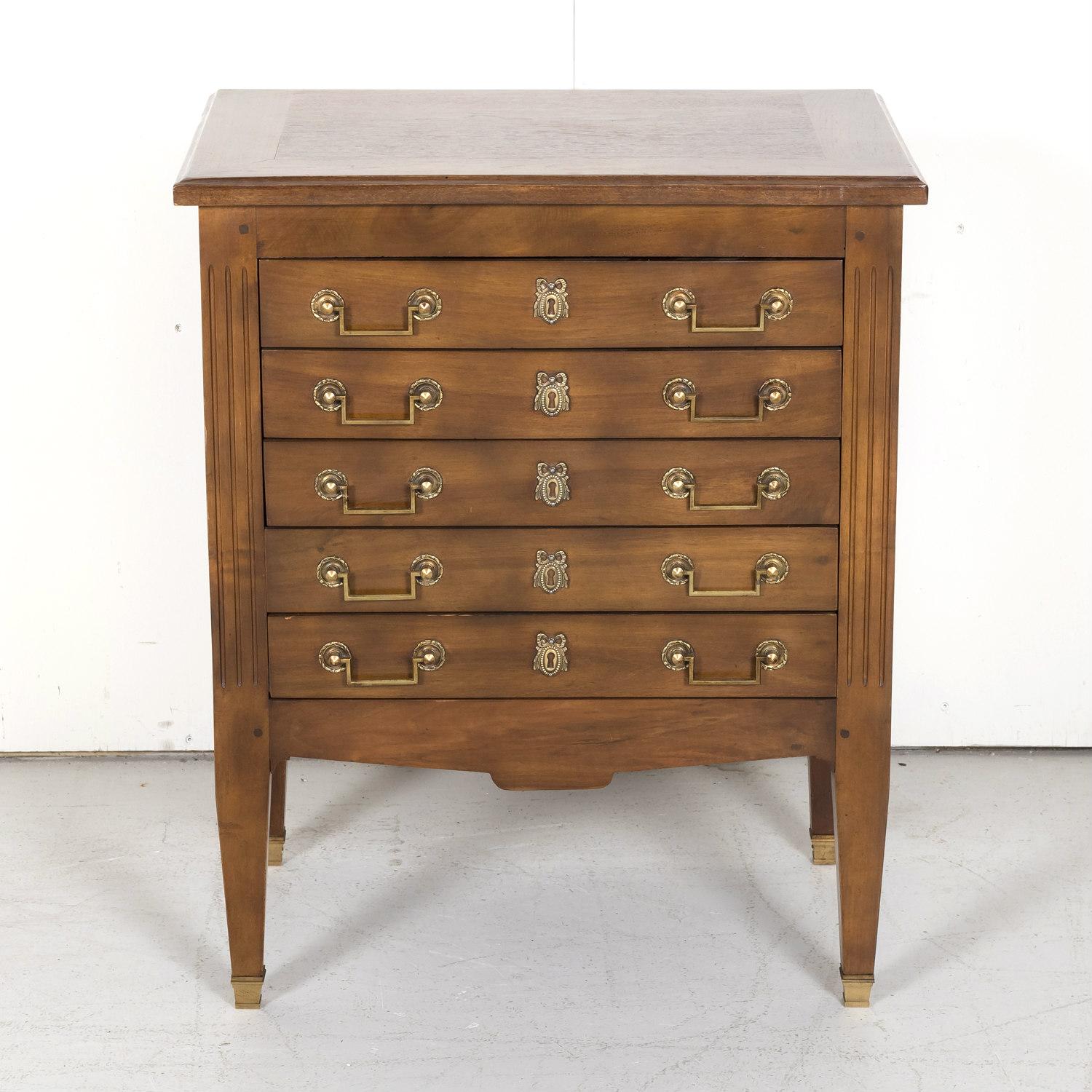 An early 20th century French Louis XVI style petite commode handcrafted of walnut having a rectangular top over five drawers with original neoclassical drop bail brass handles and brass ribbon escutcheons, circa 1900. Recessed side panels adorned
