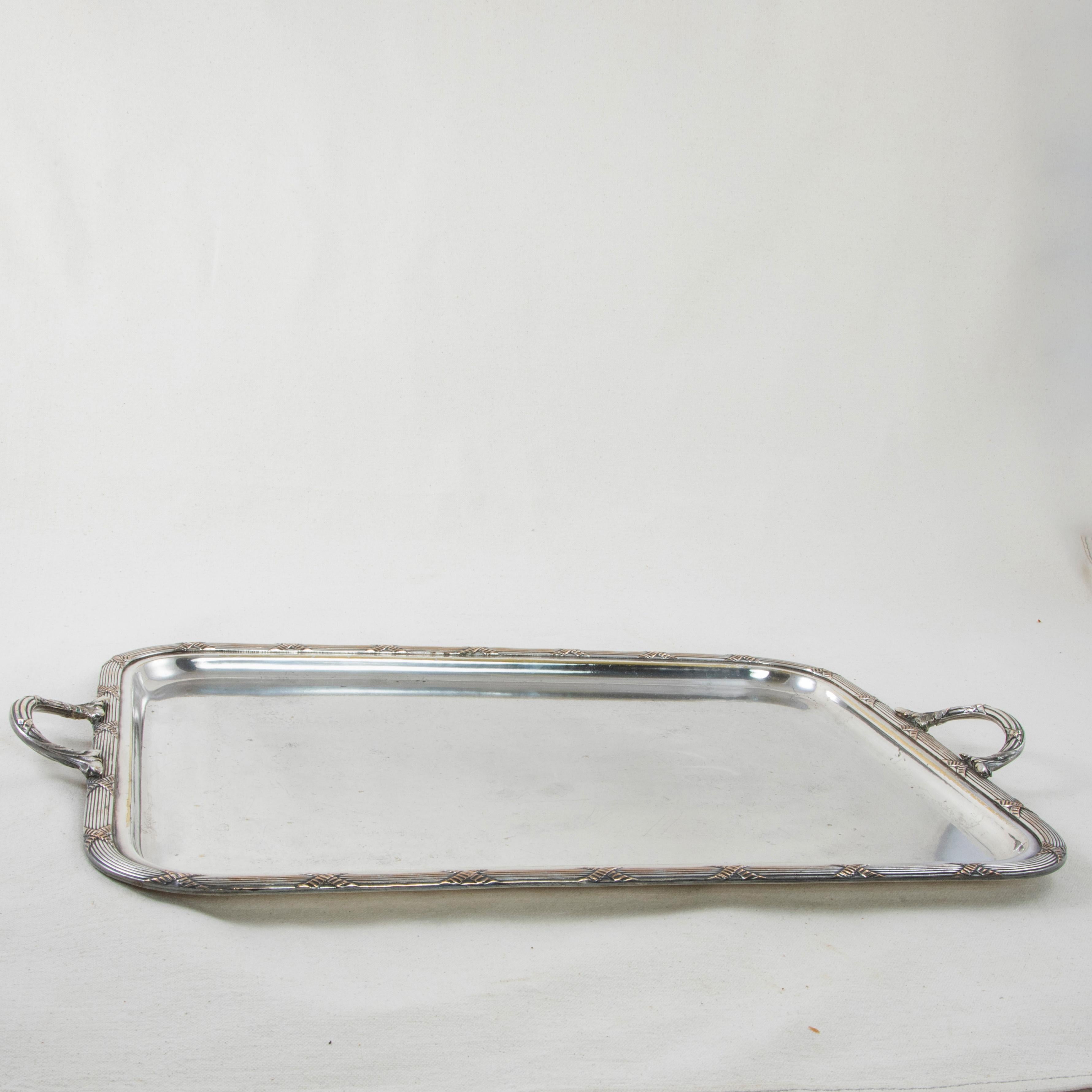 This early 20th century French silver plate serving tray features a raised rim with a classic Louis XVI motif of crossed ribbons. A handle at each end repeats the crossed ribbon motif with stylized leaves at the joints. The tray has three hallmarks