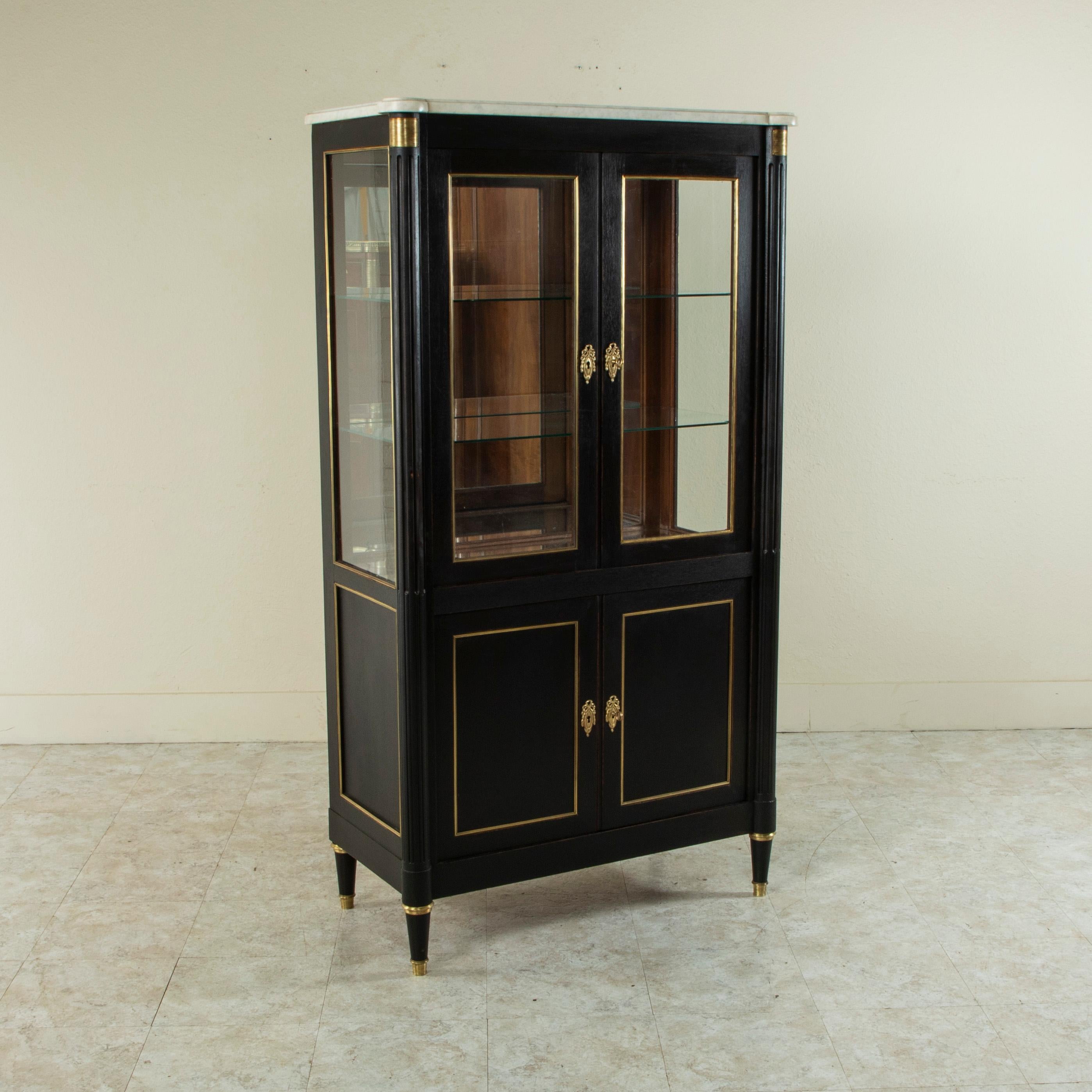This early twentieth century French Louis XVI style painted black vitrine features glass on three sides and a beveled white marble top. Two upper glass doors open to reveal an interior with two glass shelves. The back and bottom platform of the
