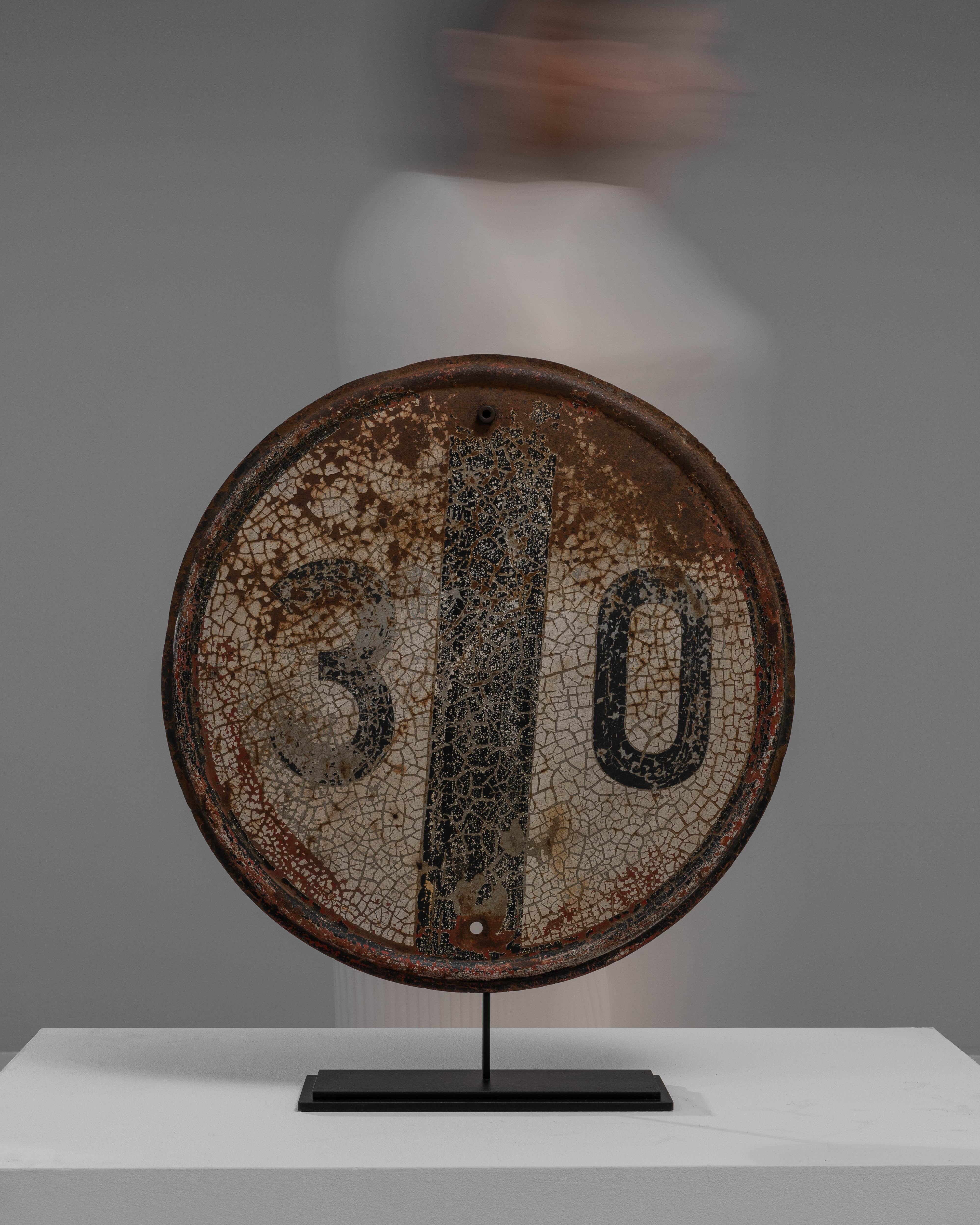Immerse yourself in the allure of antiquity with this captivating Early 20th Century French Metal Decoration on Stand. A once-functional piece of signage, this circular metal plate carries the charm of a seasoned relic, now repurposed as a
