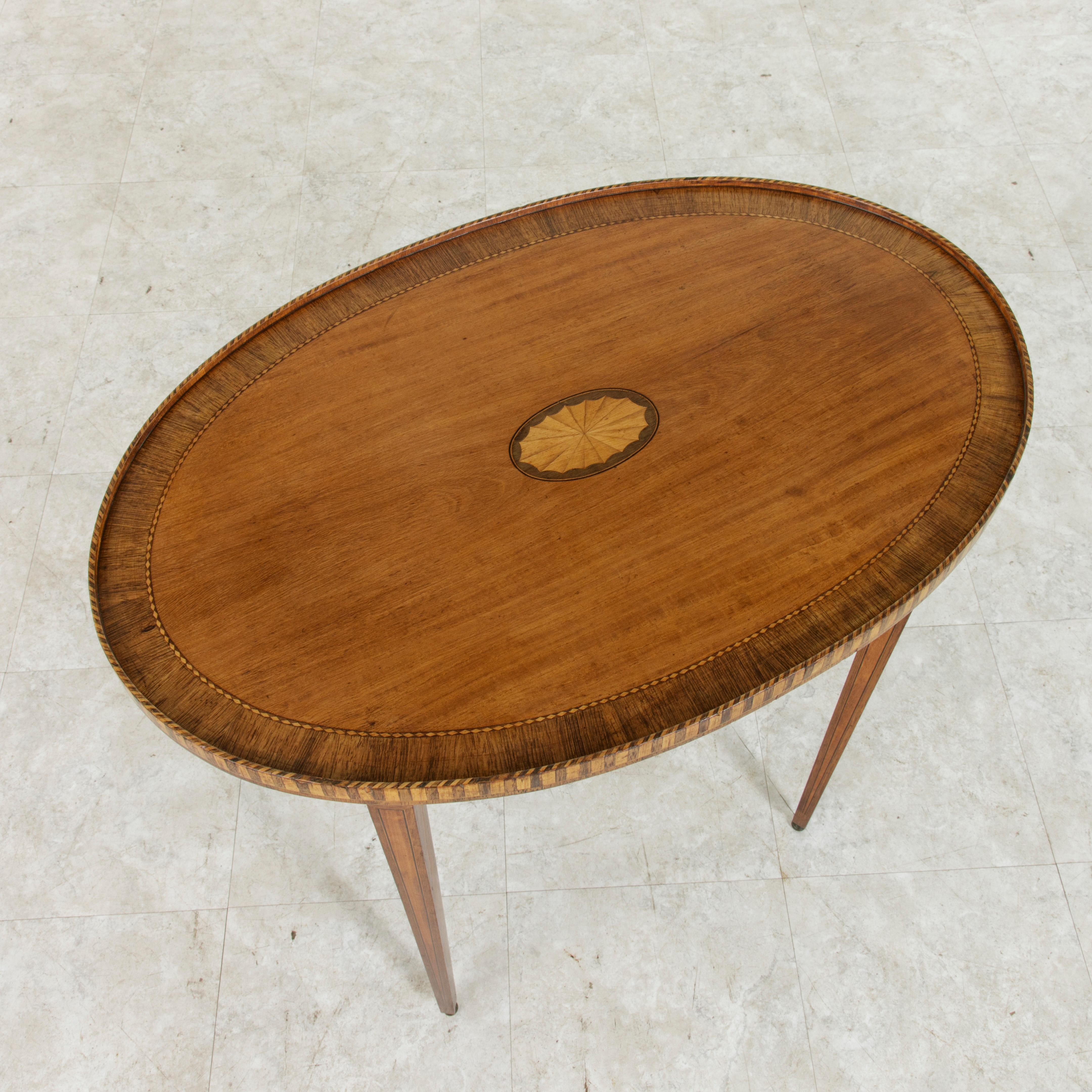 This early 20th century French neoclassic oval walnut marquetry side table features a striped rim around the edge and an inset diamond pattern border. Fine lines of inlaid lemon wood and ebonized pear wood frame the apron and tapered square legs