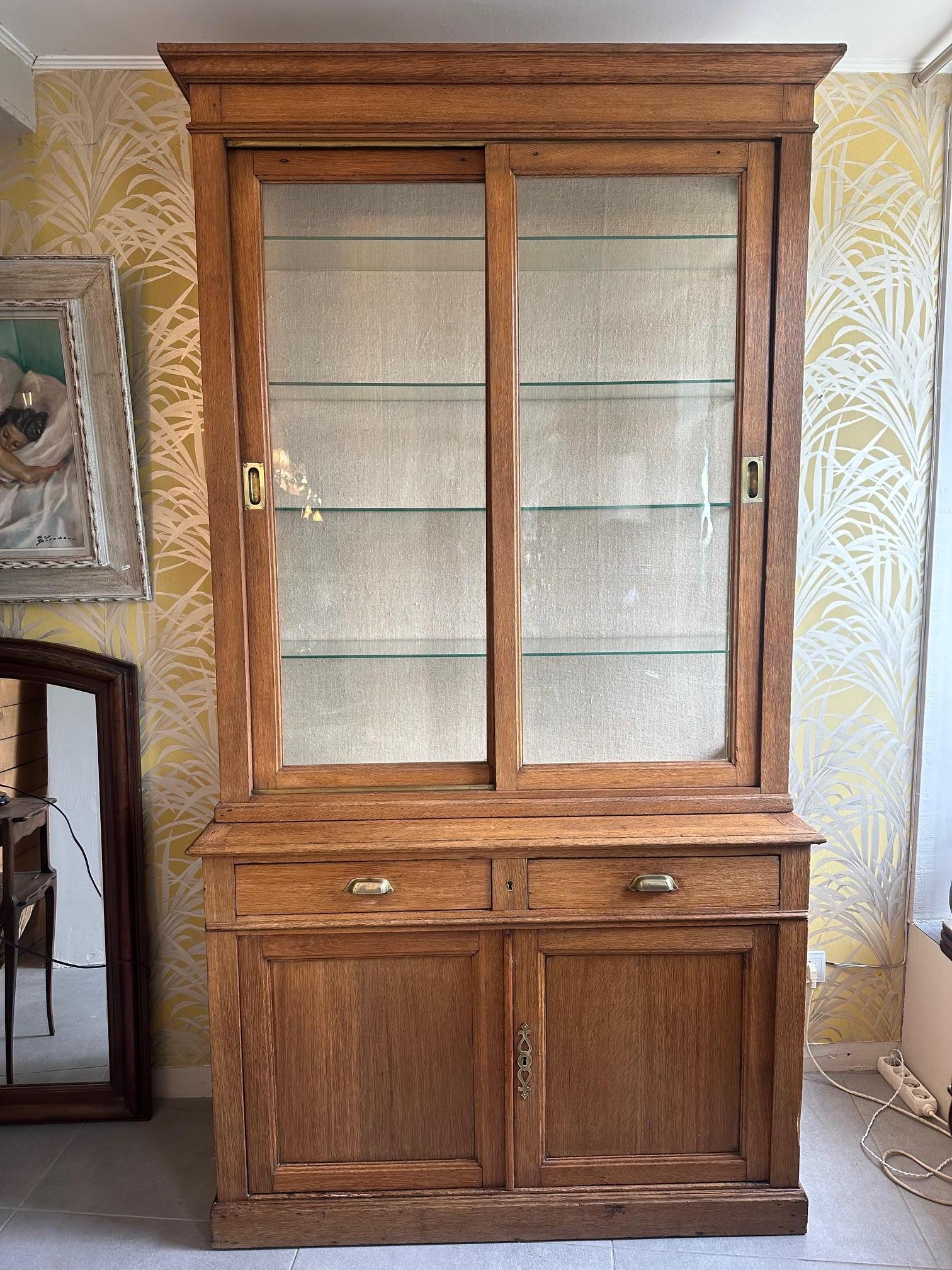 Very nice oak display cabinet dating from 1900s in good condition. 
It has two parts. The upper part has sliding glass doors and brass handles and rack. Removable glass shelves. The interior is covered in a beige linen-like fabric.
The lower part