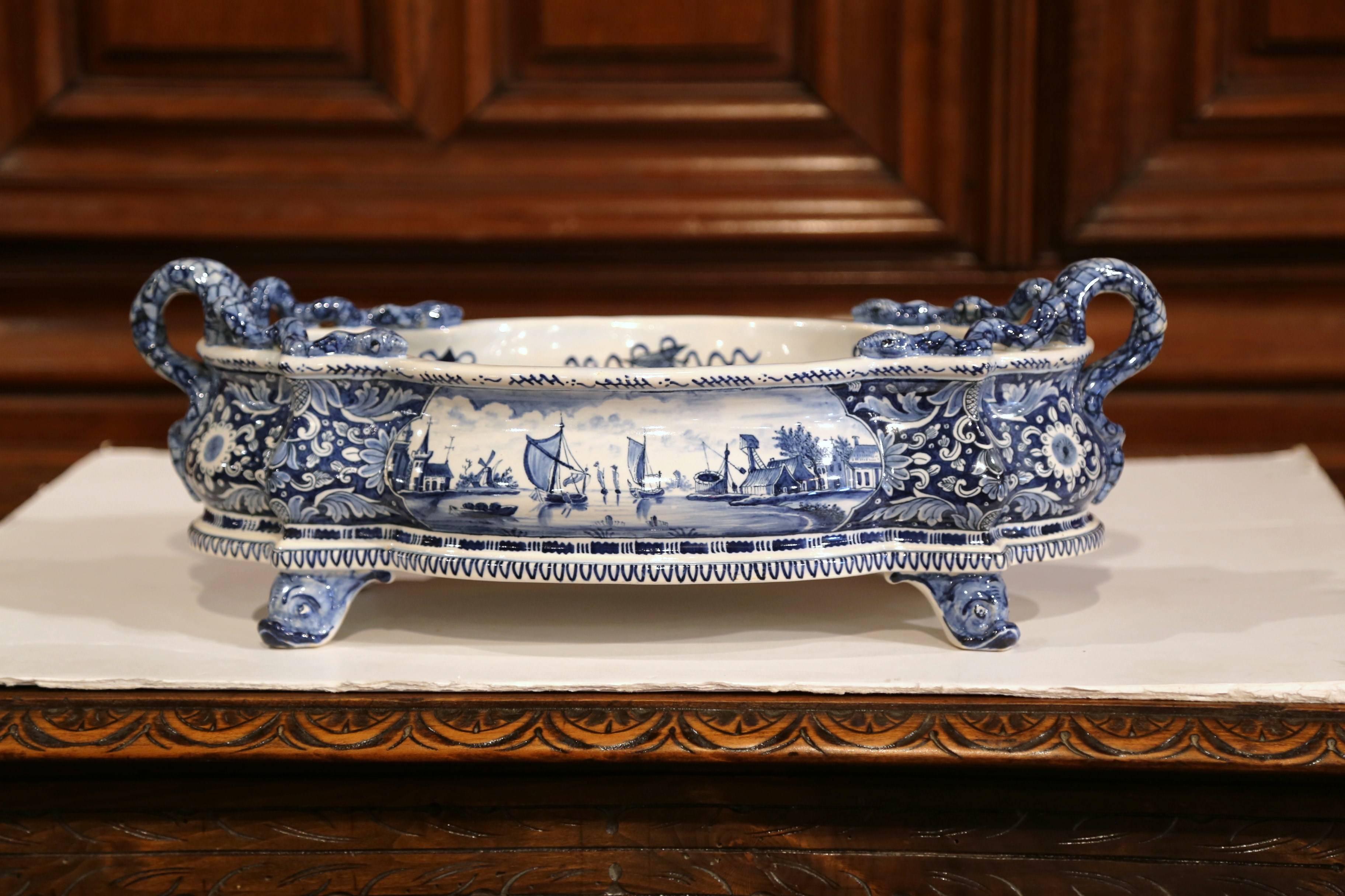 This large antique planter was crafted in France, circa 1920, oval and bowed all around, the blue and white faience jardinière features snake-shape handles, dolphin feet and hand painted medallions marine scenes in the manner of Delft with