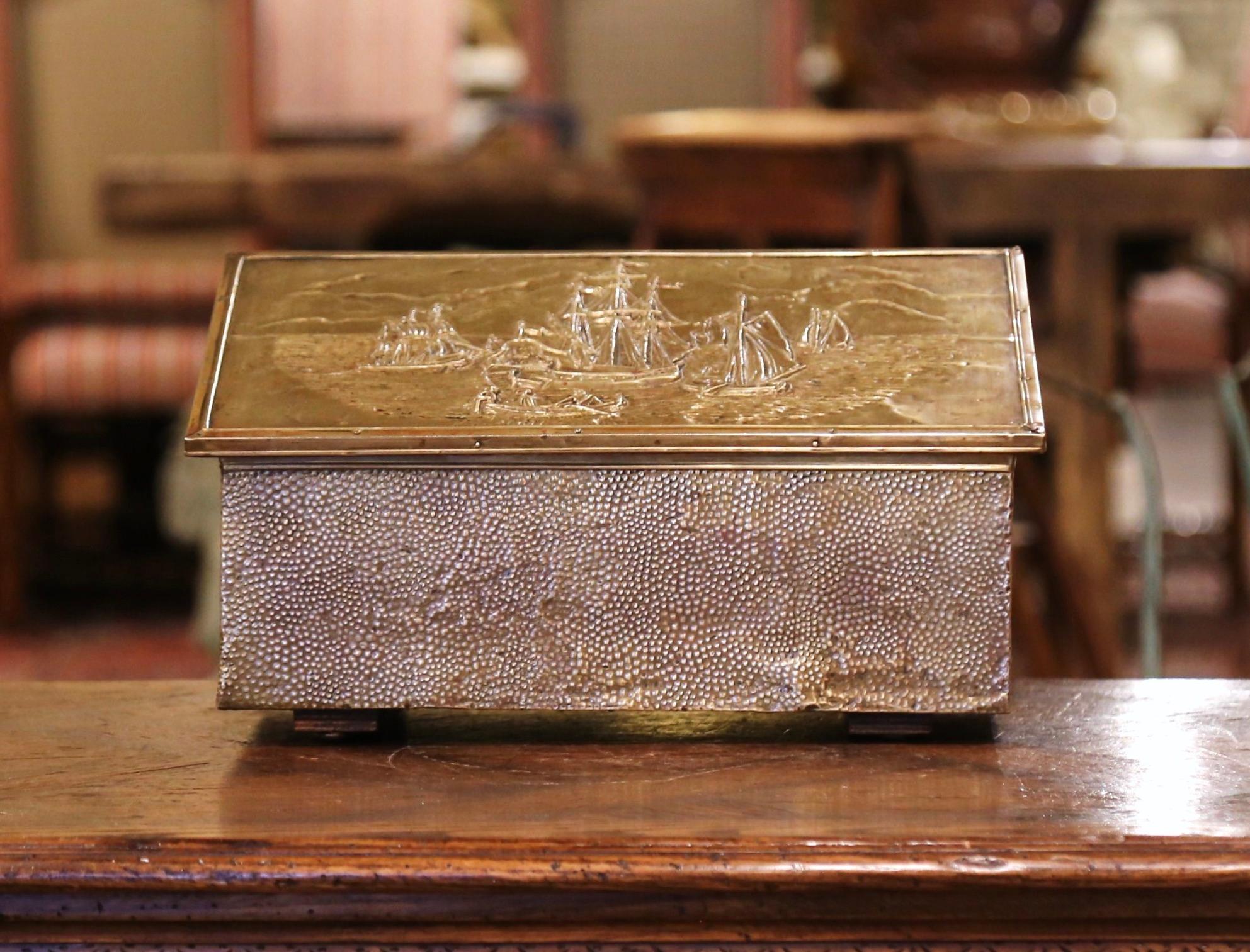 Crafted in France, circa 1920, the antique wood and brass coffer is decorated on the top with a repousse sail boat scene in the manner of Claude Vernet. The slant top opens to inside storage upholstered with green velvet. The small decorative trunk