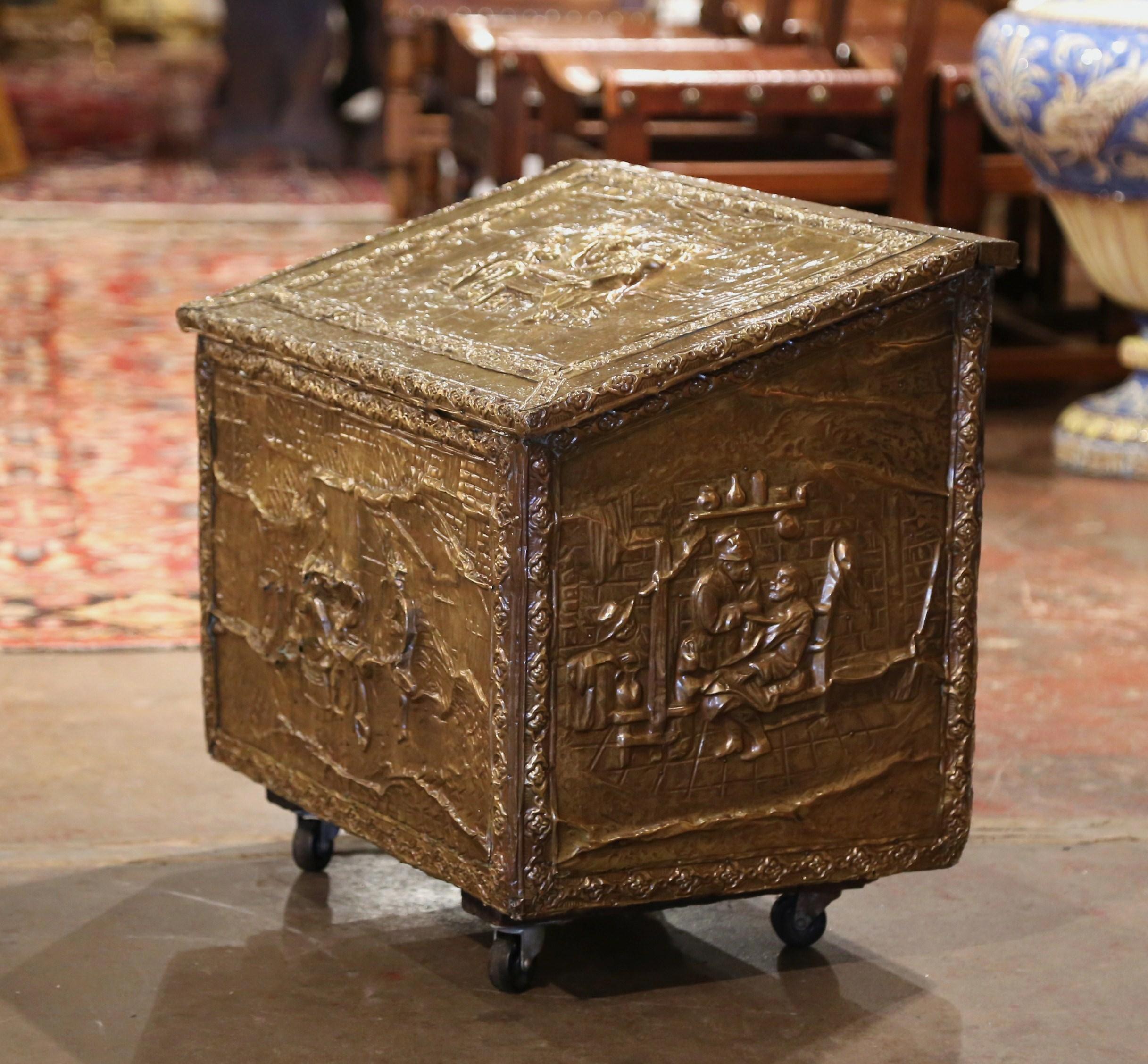 Crafted in France, circa 1920, the antique wood and brass fireplace wood coffer stands on casters; the trunk is decorated with repousse tavern scene motifs in the manner of David Teniers on all four sides including the top and front; each scene