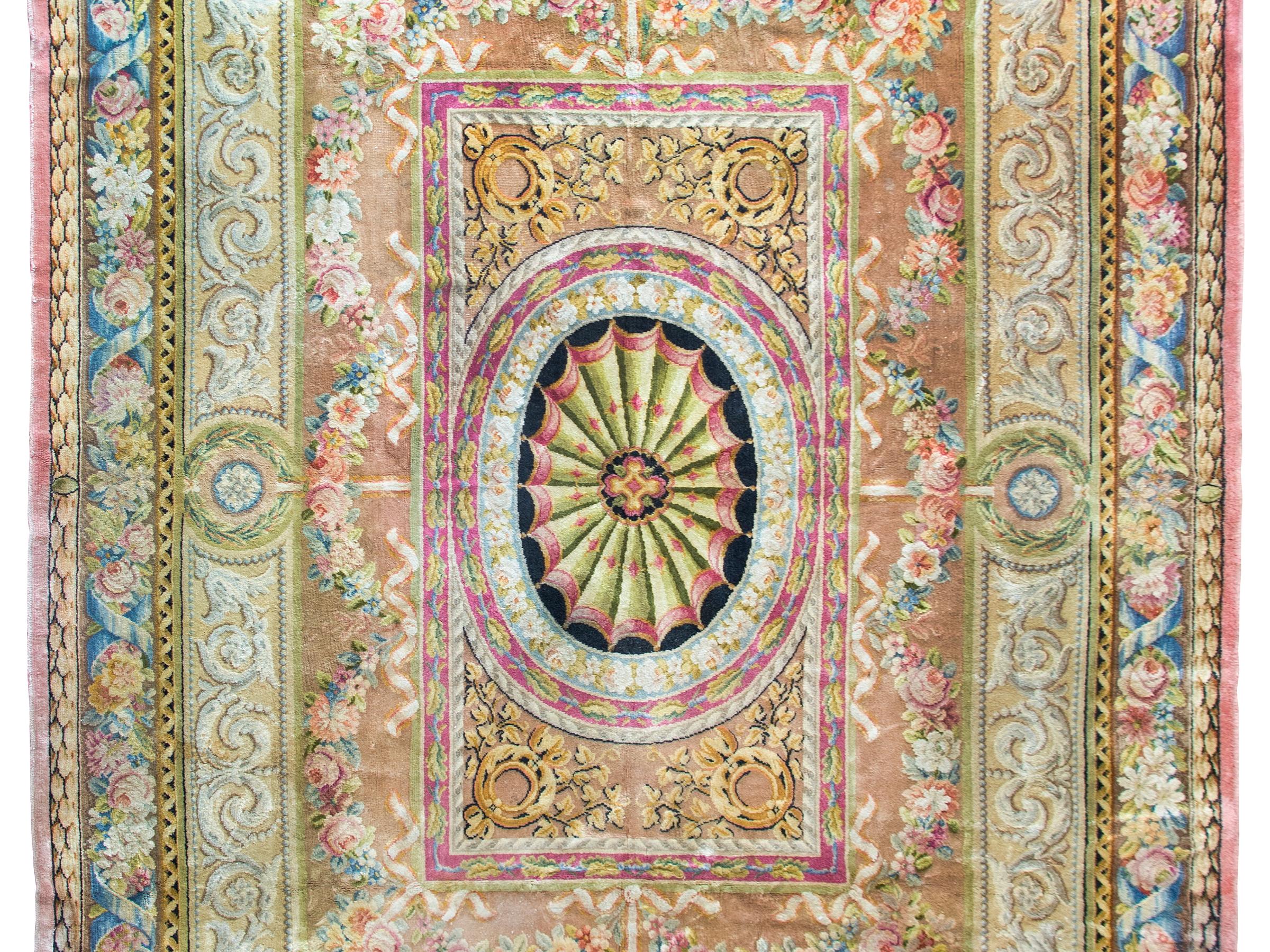 This exquisite early 20th-century French Savonnerie rug showcases an elaborate pattern rich with large-scale floral swags, acanthus leaves, ribbon twists, and a variety of neoclassical motifs. Woven in a vibrant palette of brilliant pinks, indigos,