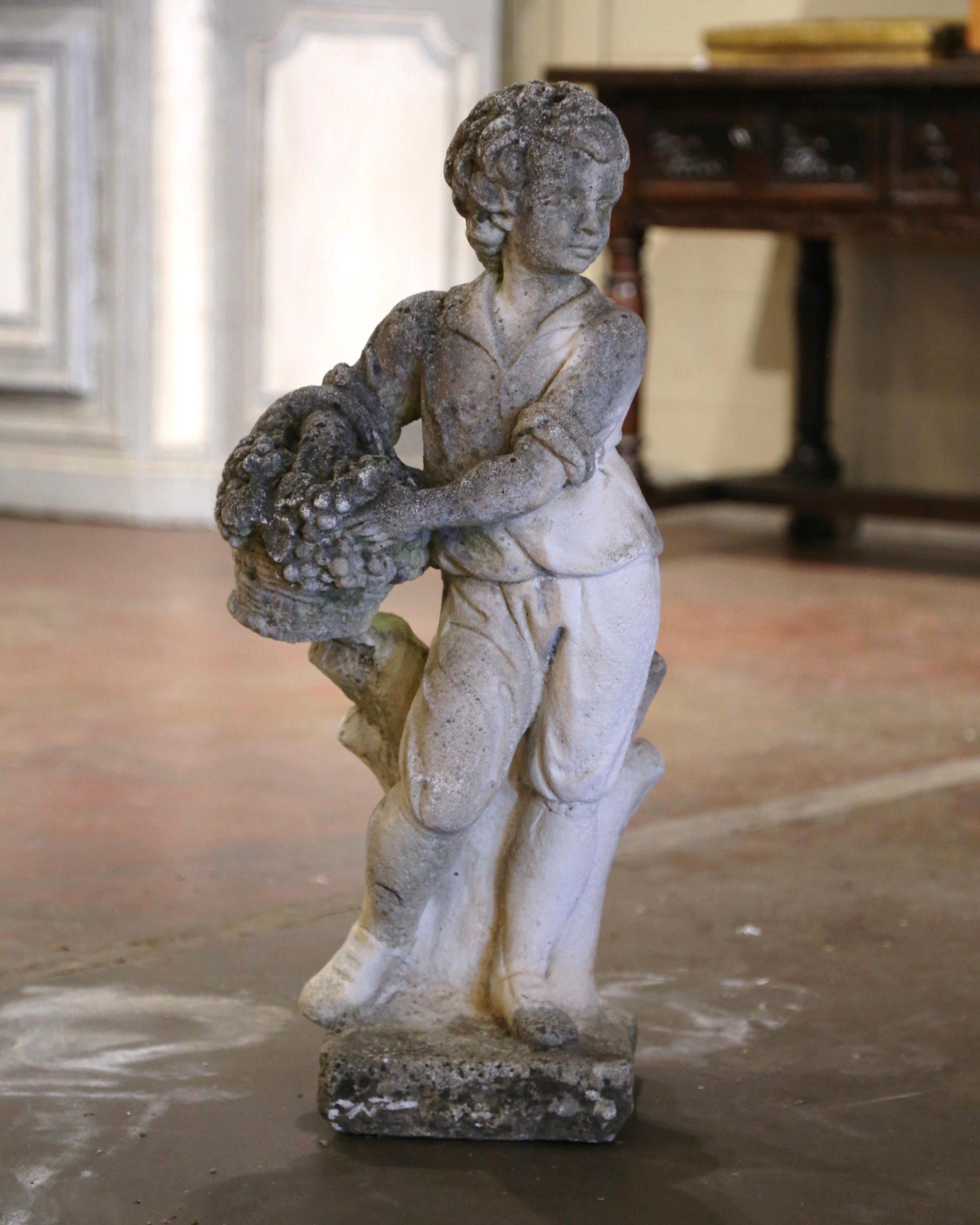 Decorate a garden or patio with this elegant antique outdoor statue. Made of cast stone in France circa 1920, the tall sculpture depicts a youngster standing on a square pedestal, holding a basket full of grapes, and dressed in a tunic, with one leg