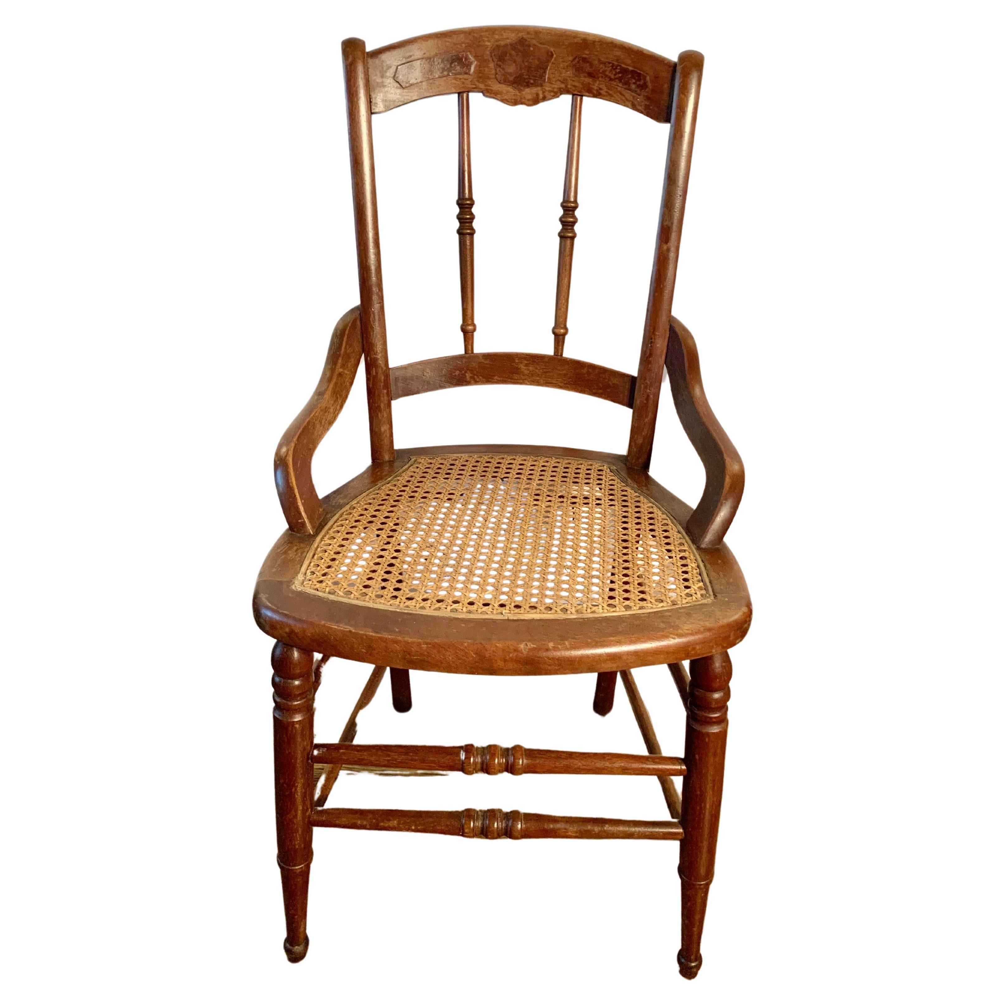 Cette chaise du début du XXe siècle a été fabriquée en France en noyer avec une assise en rotin au début des années 1900. La chaise est dotée d'un dossier en fuseau et d'une traverse supérieure sculptée à la main d'un blason familial. La pièce