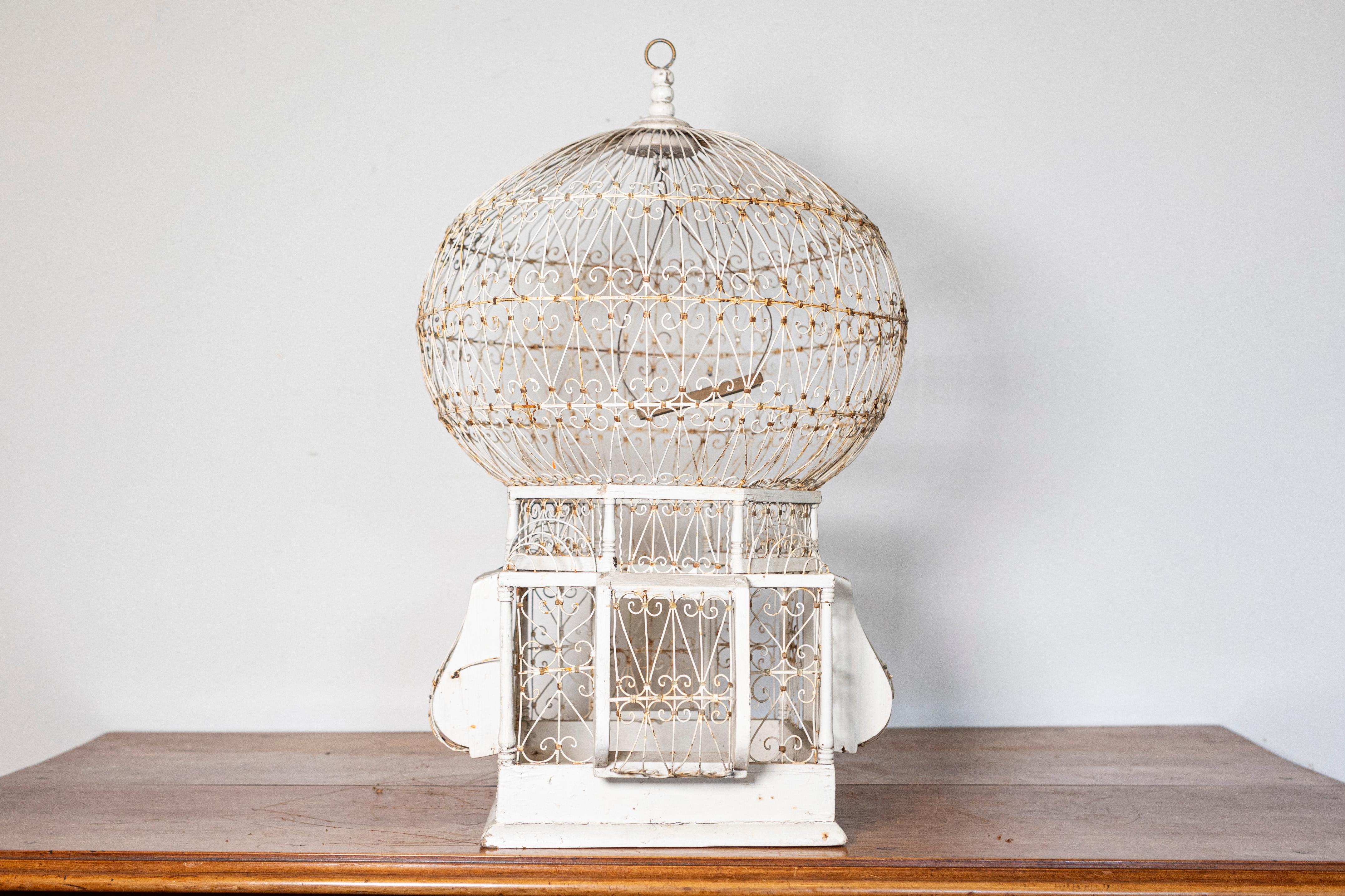 This exquisite early 20th-century French birdcage showcases a beautifully crafted white-painted wrought iron structure with an elegant domed top, embodying the delicate craftsmanship of the period. The intricate scrollwork detailing, combined with
