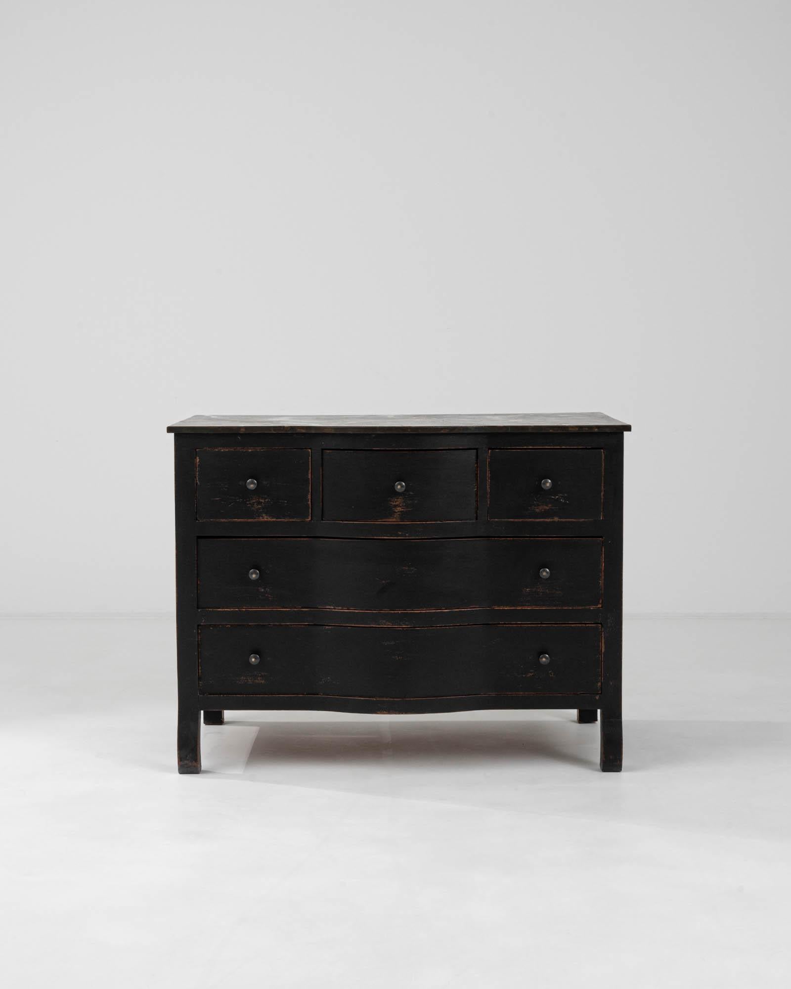 This early 20th Century French wood chest of drawers exudes a rich sense of history with its black patinated finish, offering a striking statement piece for any room. The chest features a classic design with elegant curves and a stately stance,
